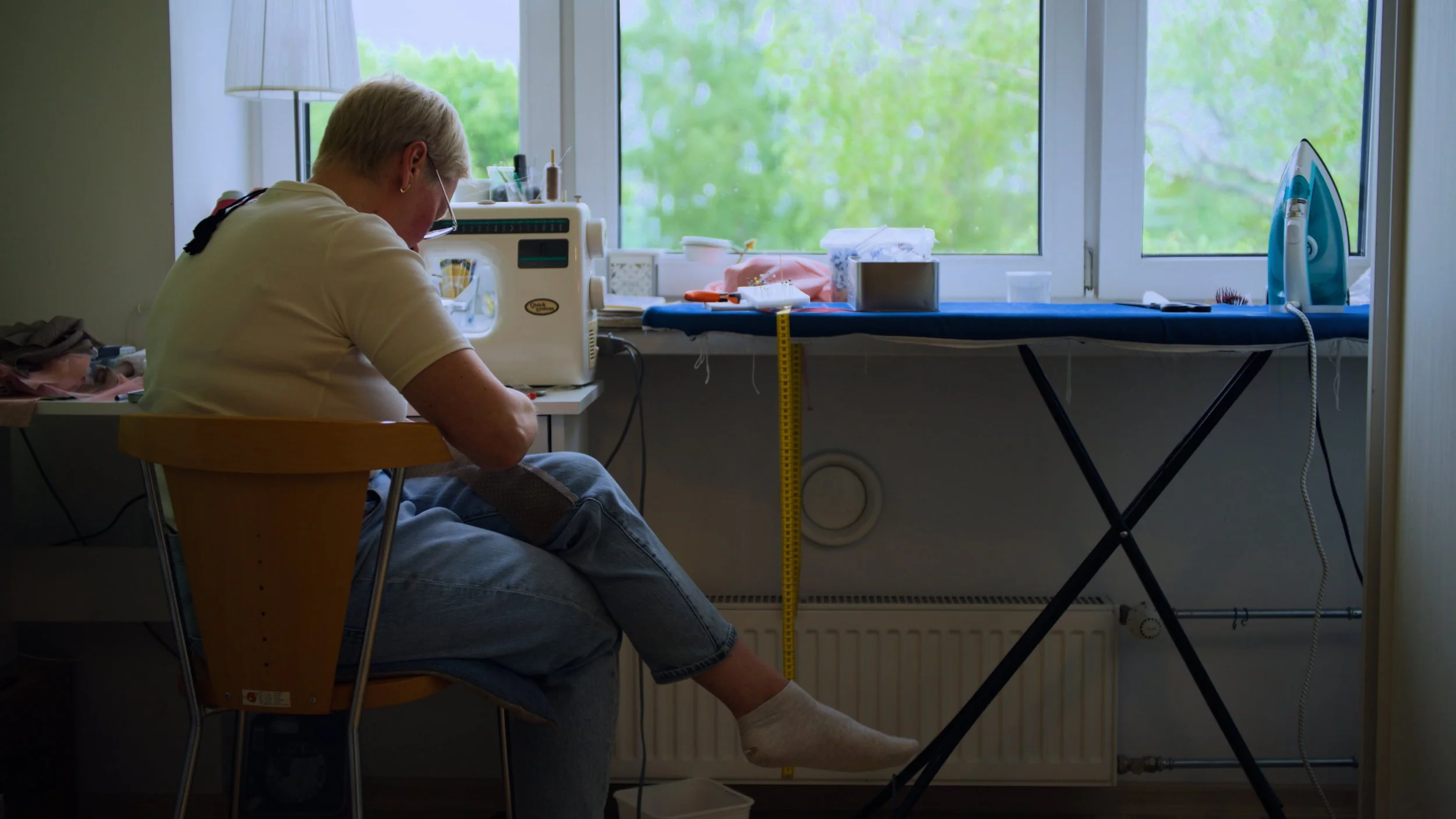 Person sitting on a chair at a desk working with a sewing machine, in front of a window with green trees outside, and an iron on an ironing board to the right.