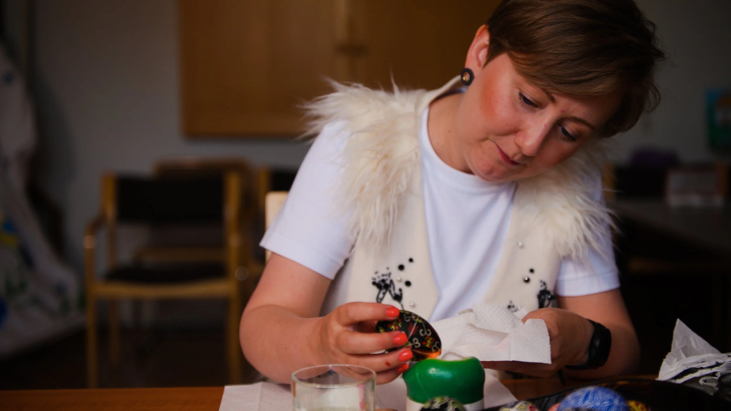 A woman with short brown hair wearing a white shirt with a faux fur vest decorating colorful Ukrainian pysanka Easter eggs at a table.