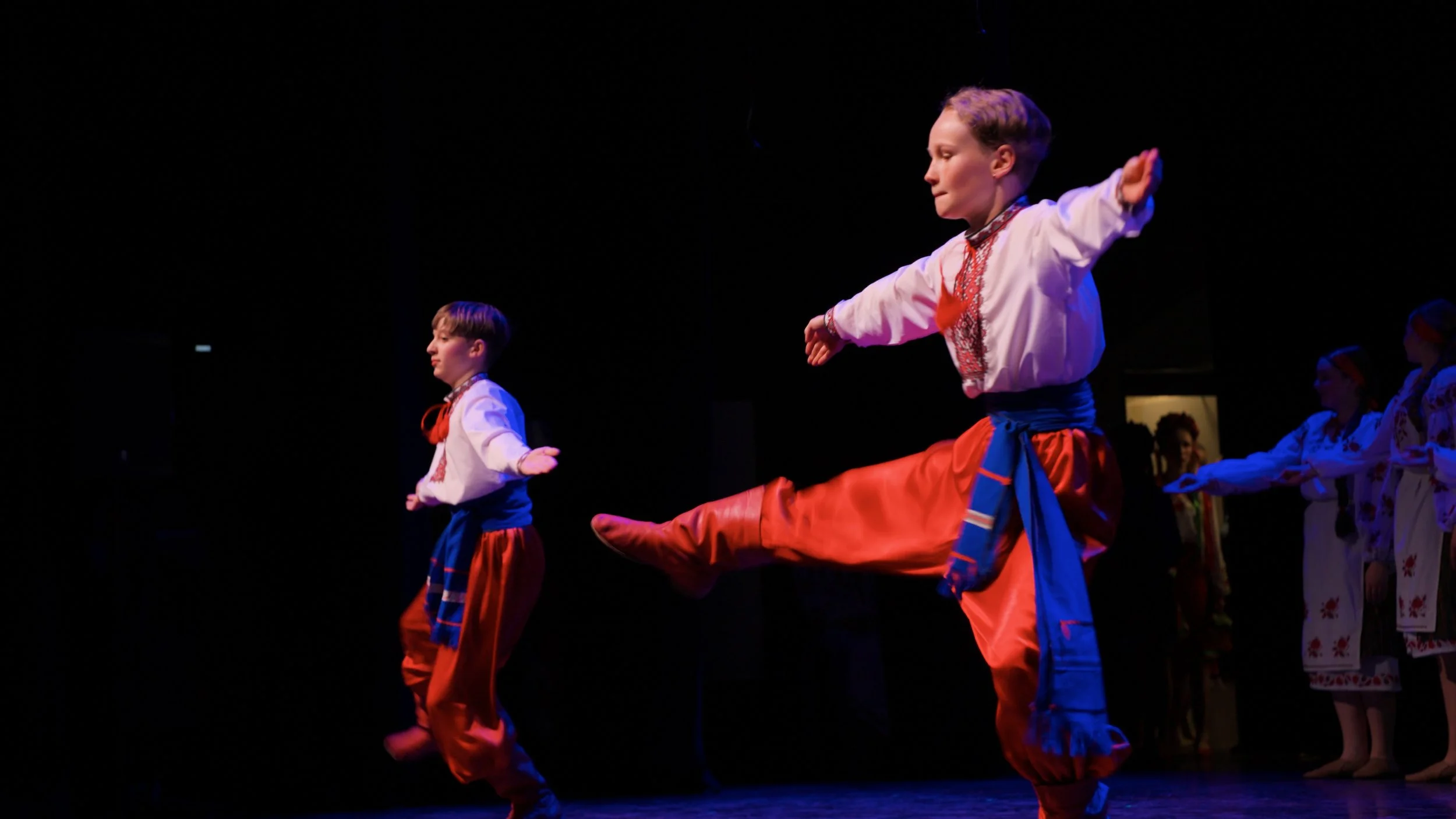 Children dressed in traditional Ukrainian clothing perform a dance on stage. The boys are wearing embroidered shirts and bright pants, with one boy kicking his leg high in the air and others balancing on one foot.