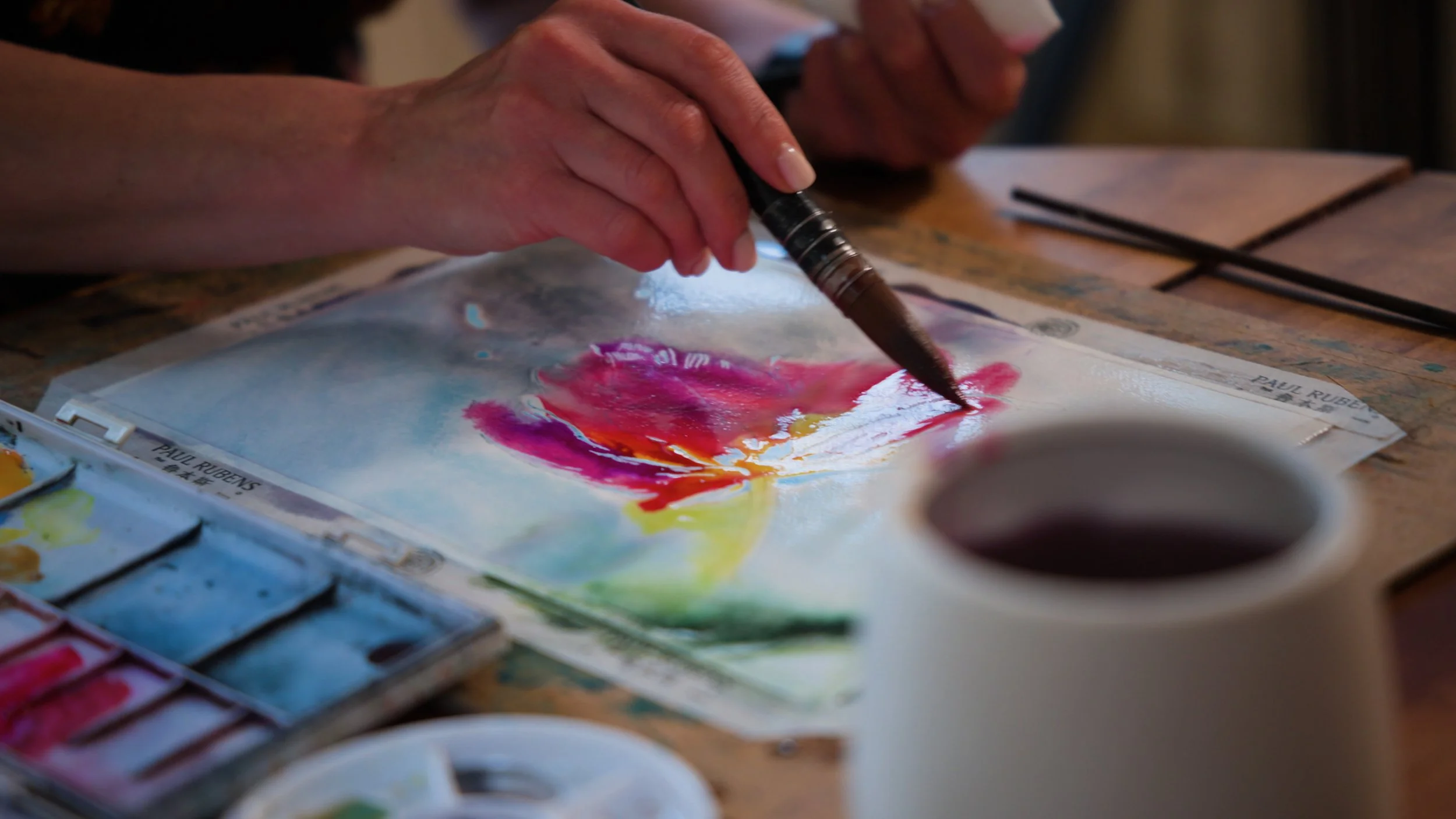 Person painting a colorful watercolor artwork of a flower on paper, with watercolor paints and brushes on the table.