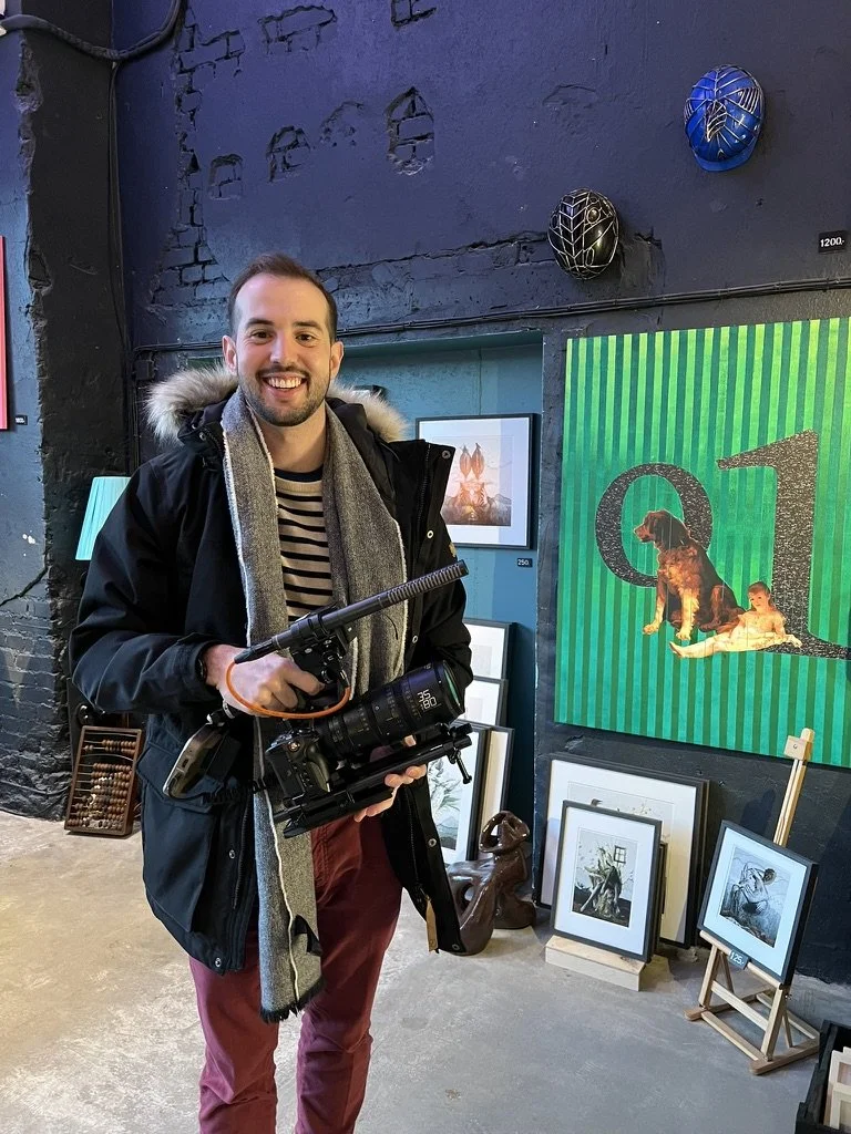 Man smiling at camera holding a professional video camera inside an art gallery with framed artwork and decorative masks on the wall.
