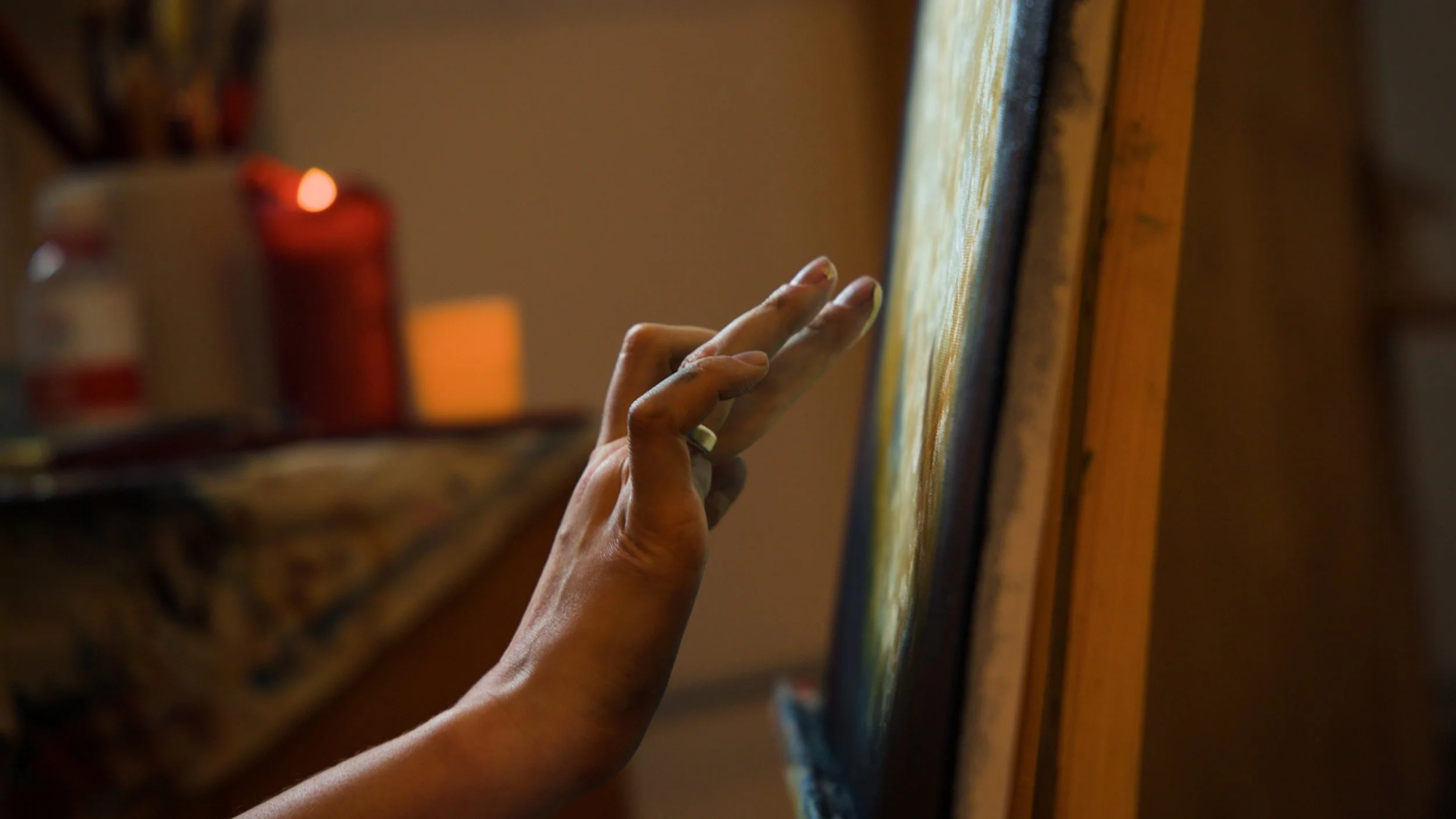 A person's hand touching a textured painting on a canvas, with a blurred background featuring a red candle and a bottle.