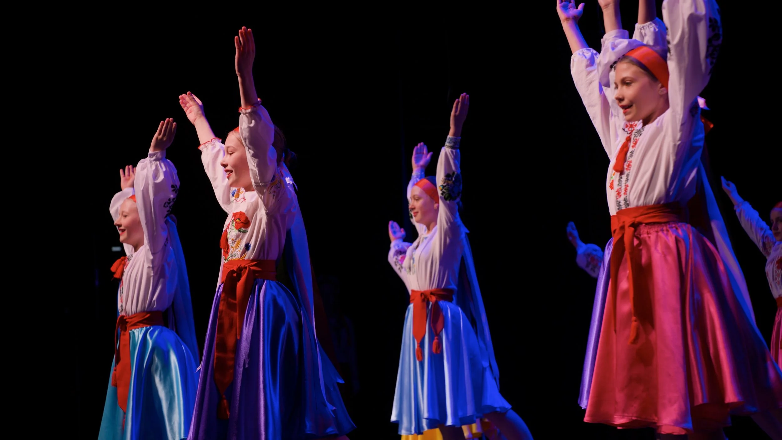 Girls in traditional Ukrainian folk costumes performing a dance on stage with arms raised.