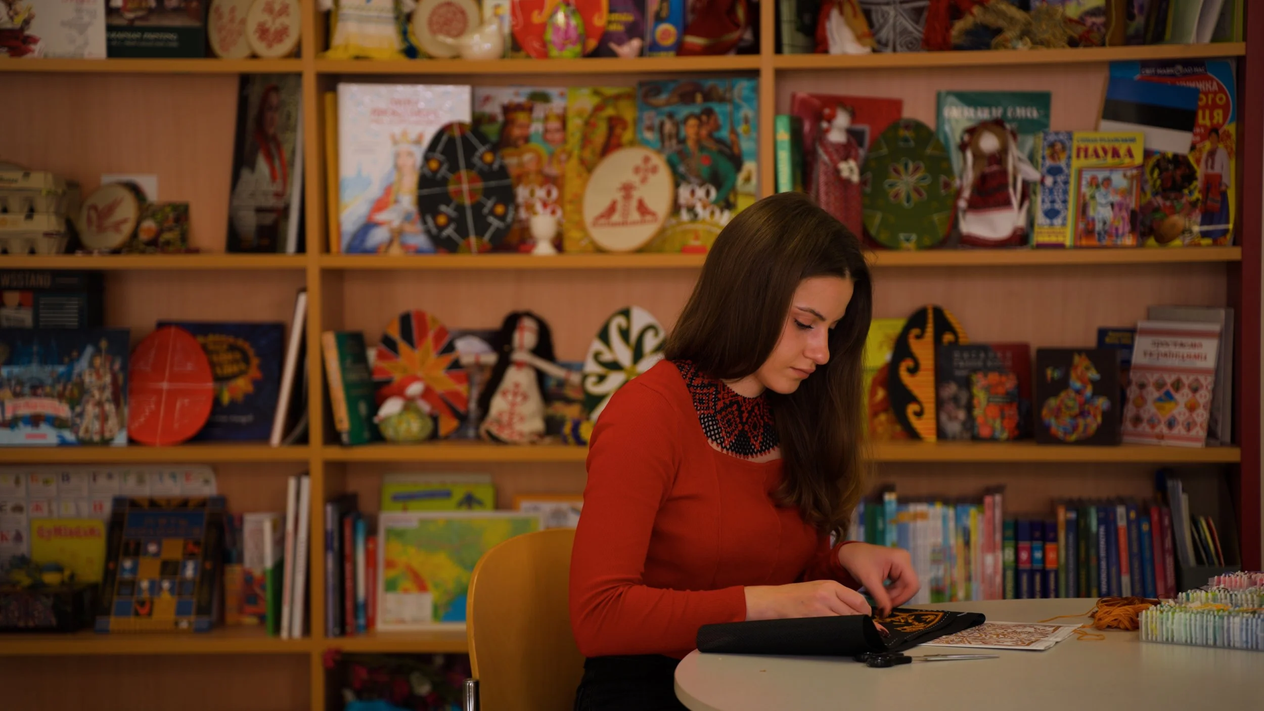 A woman sitting at a table with crafts and embroidery supplies, in front of a bookshelf filled with colorful books and decorative items.
