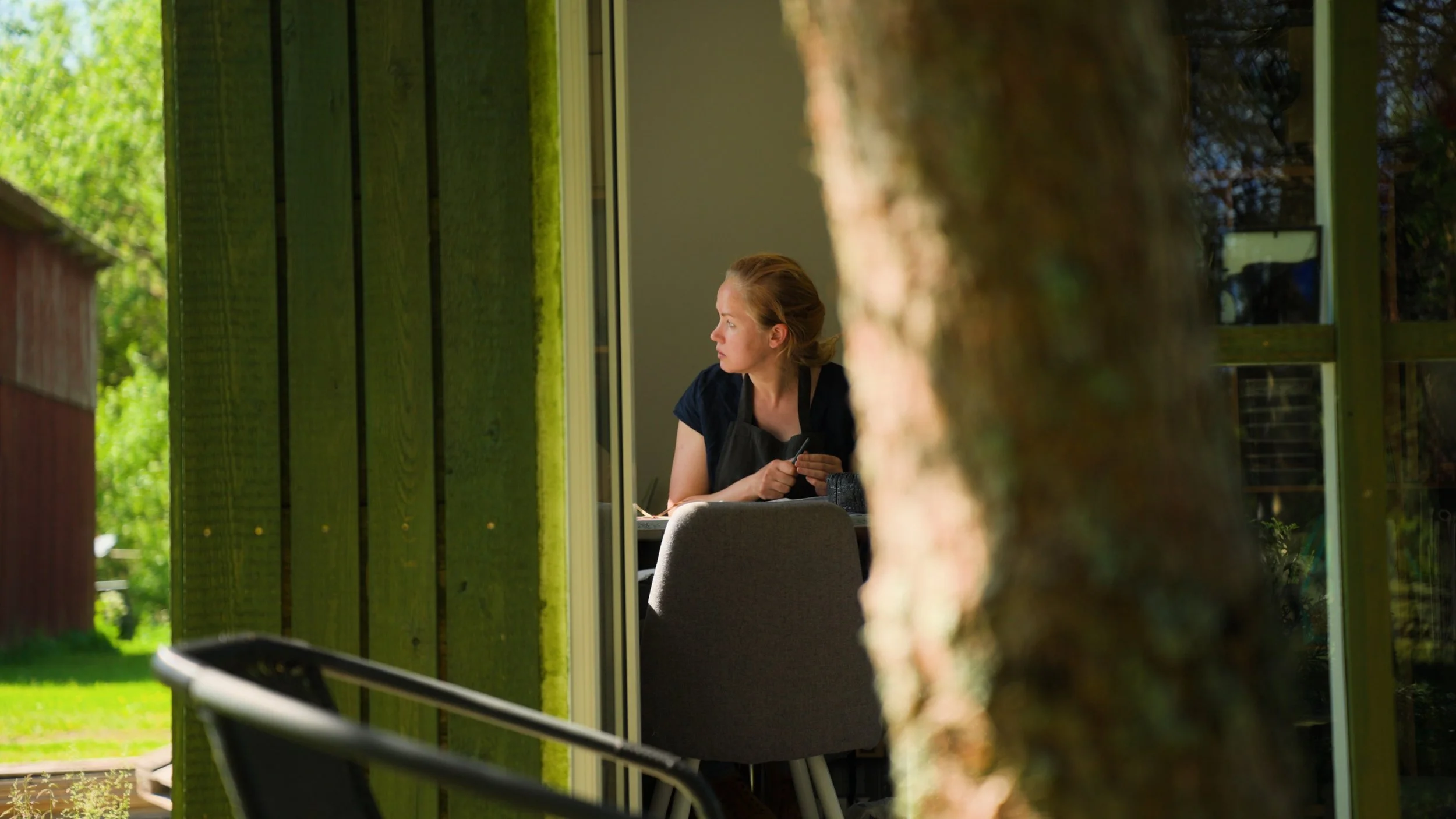 A woman sitting indoors near a window, looking outside, with a tree partially blocking the view.
