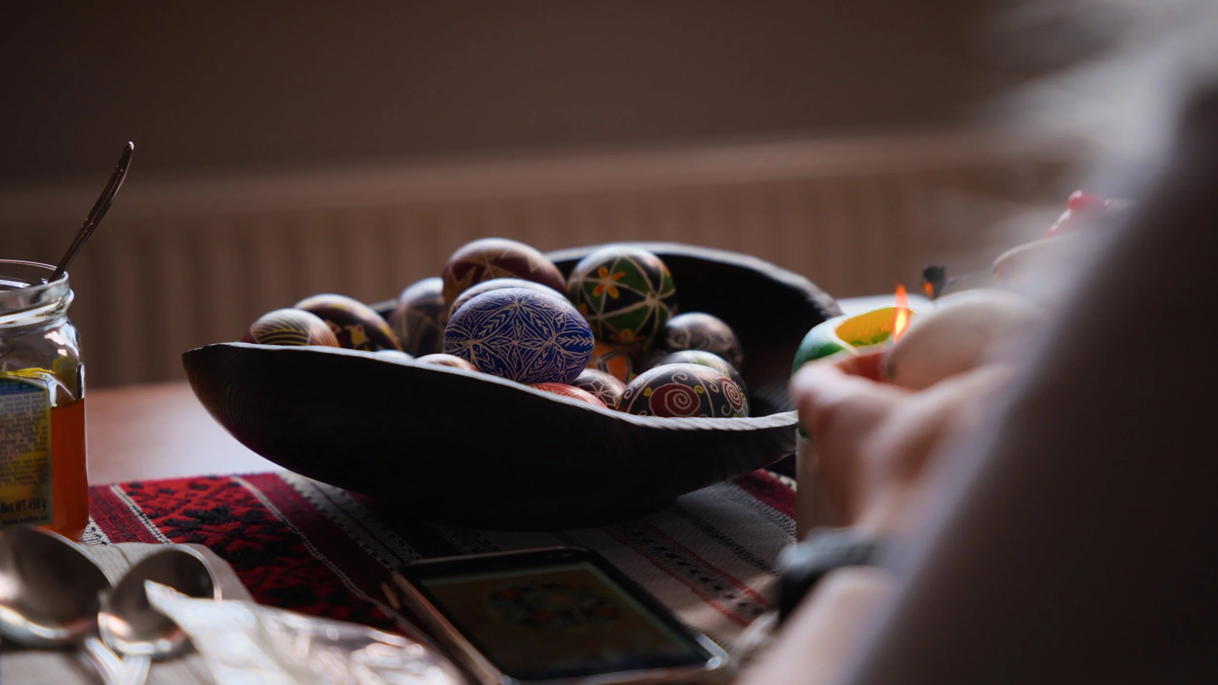 Decorated pysanka Easter eggs in a black bowl on a table with a patterned tablecloth; some eggs are on the table, including one with a smoky pattern and another with a floral design.
