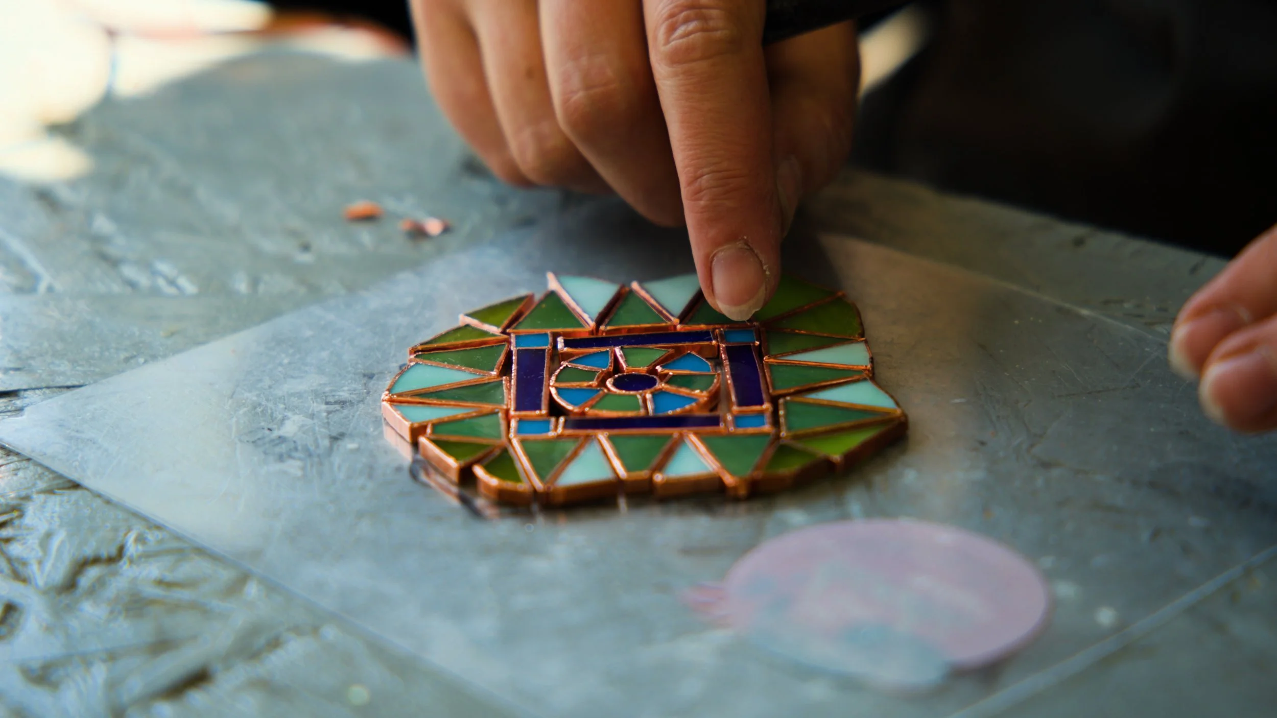 A person is working on a stained glass piece with a colorful geometric design in the style of Ukrainian pysanka egg art, using their finger to adjust the glass pieces.