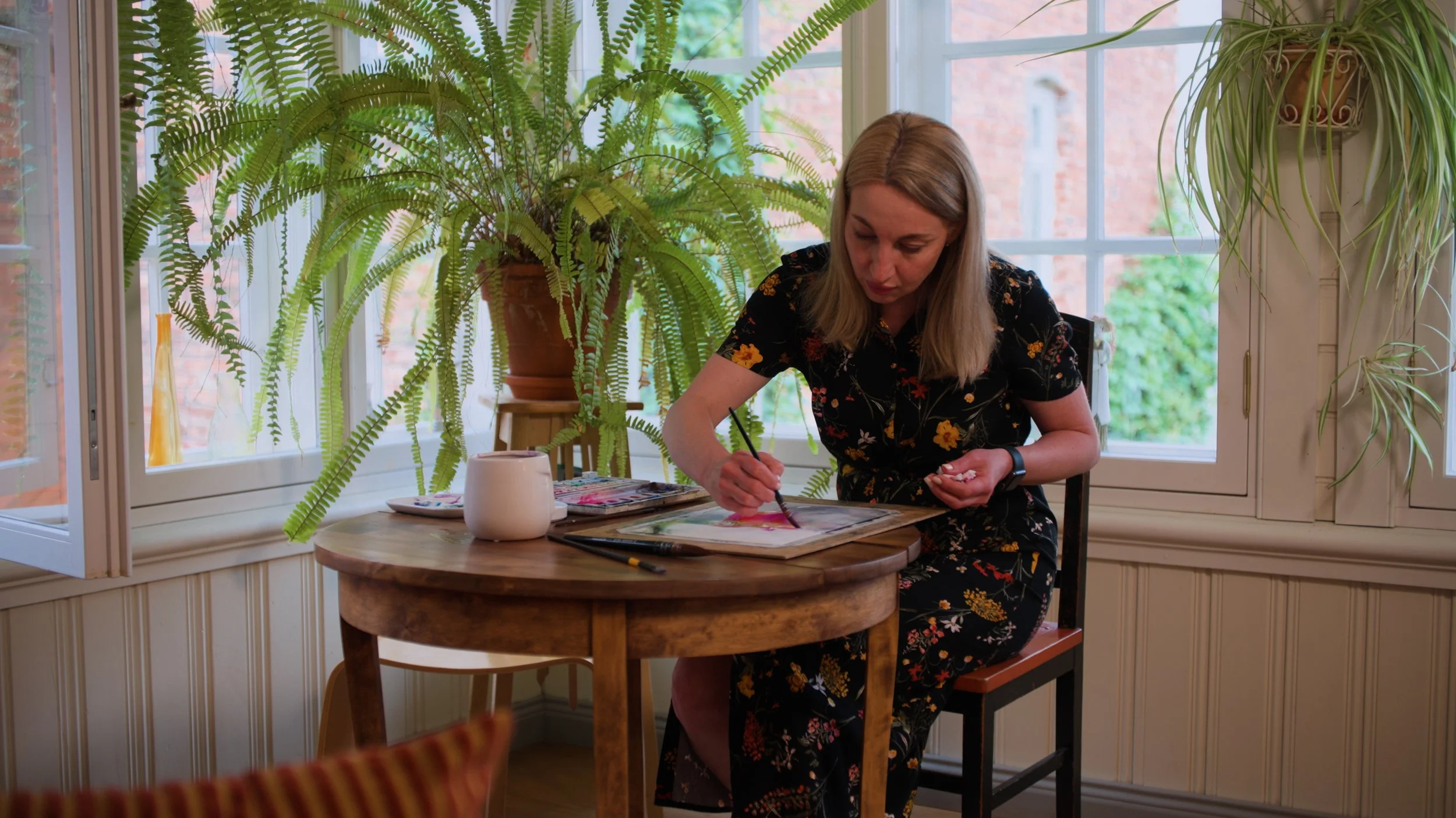 A woman painting with watercolor on paper at a wooden table in a well-lit room with large windows and green plants.