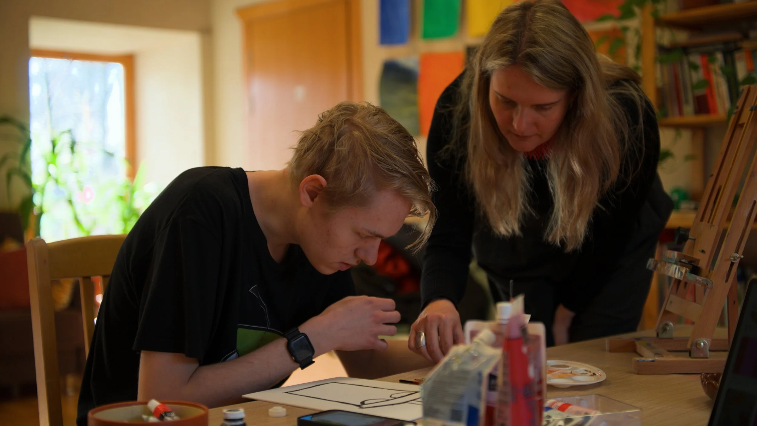 A woman guiding a young man as he paints or draws on a canvas or paper in a cozy, well-lit room with bookshelves and colorful artwork.