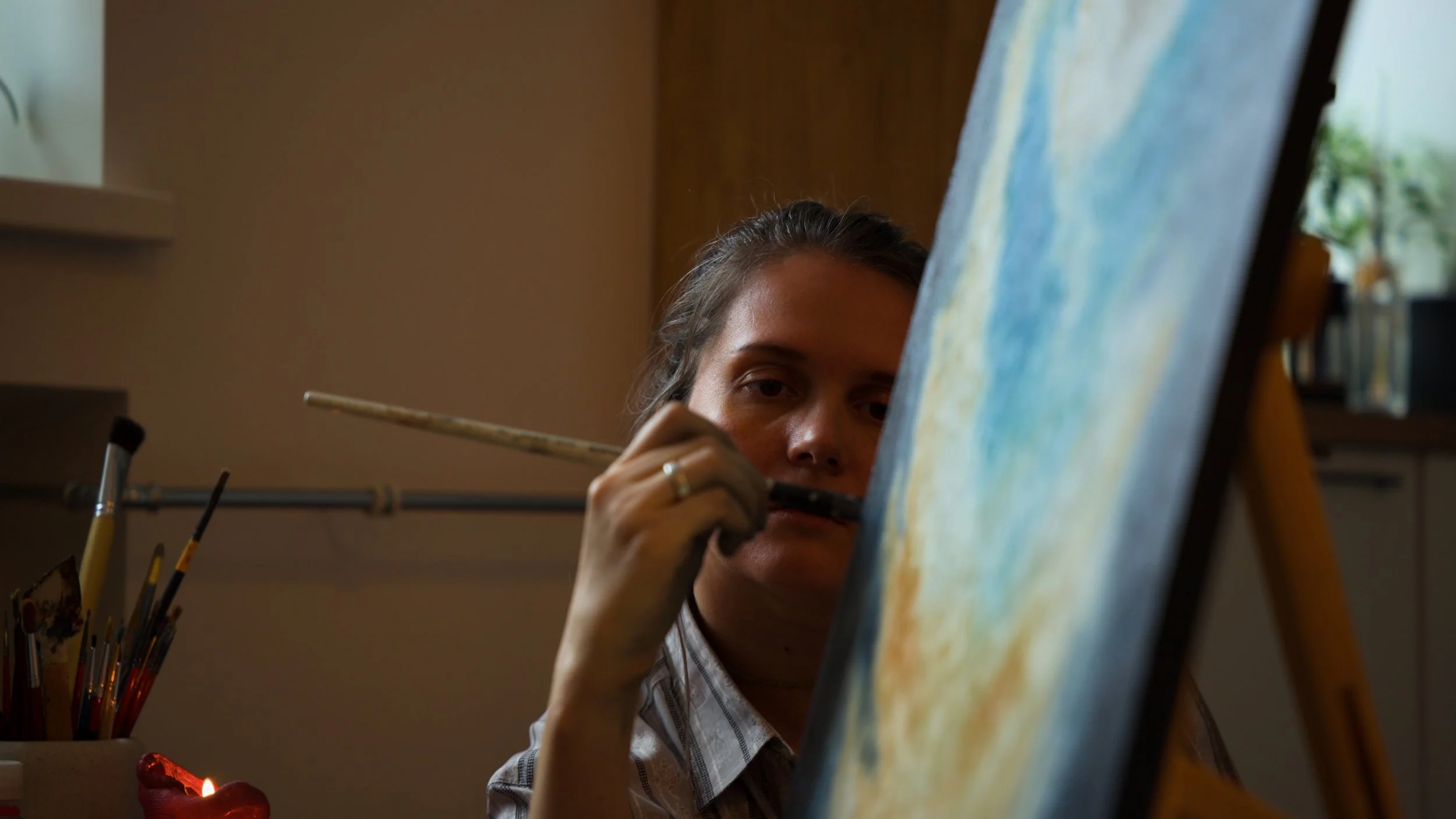 An artist is painting on a canvas placed on an easel, holding a brush close to her face, with art supplies in the background.
