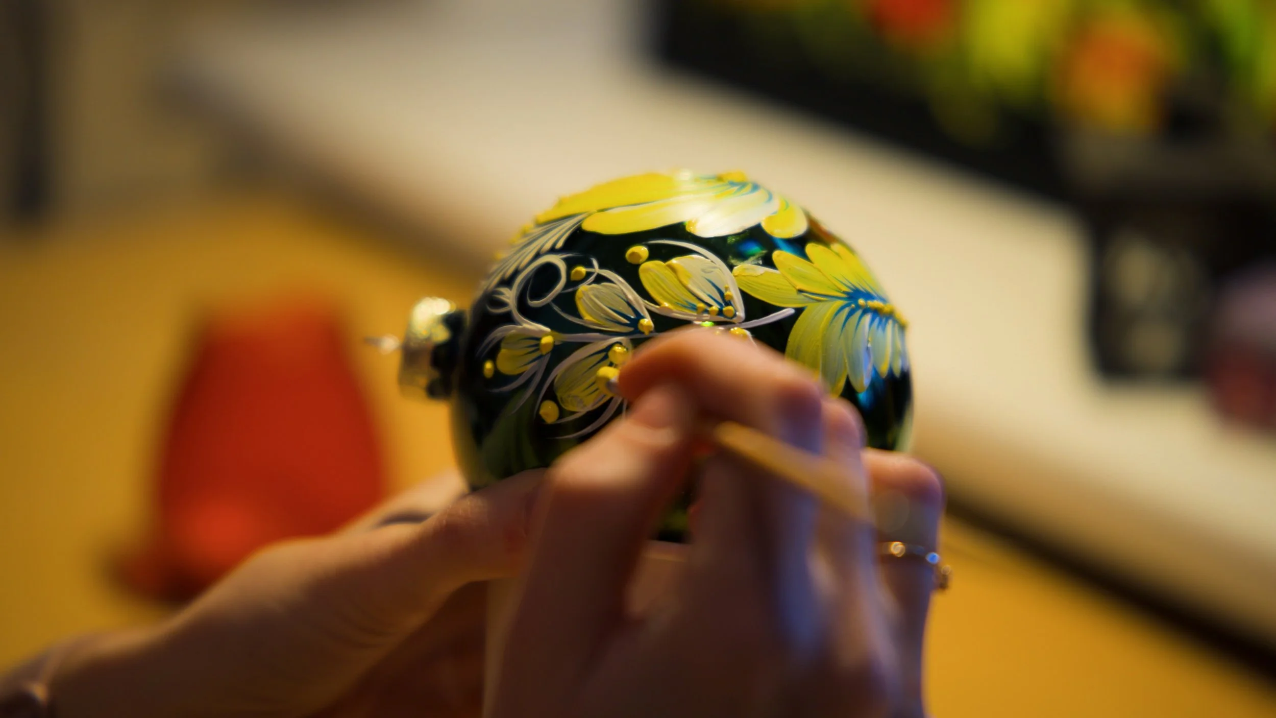 A person's hand holding a decorated black Christmas ornament with yellow and blue floral designs, painting it in the style of Ukrainian Petrykivka.