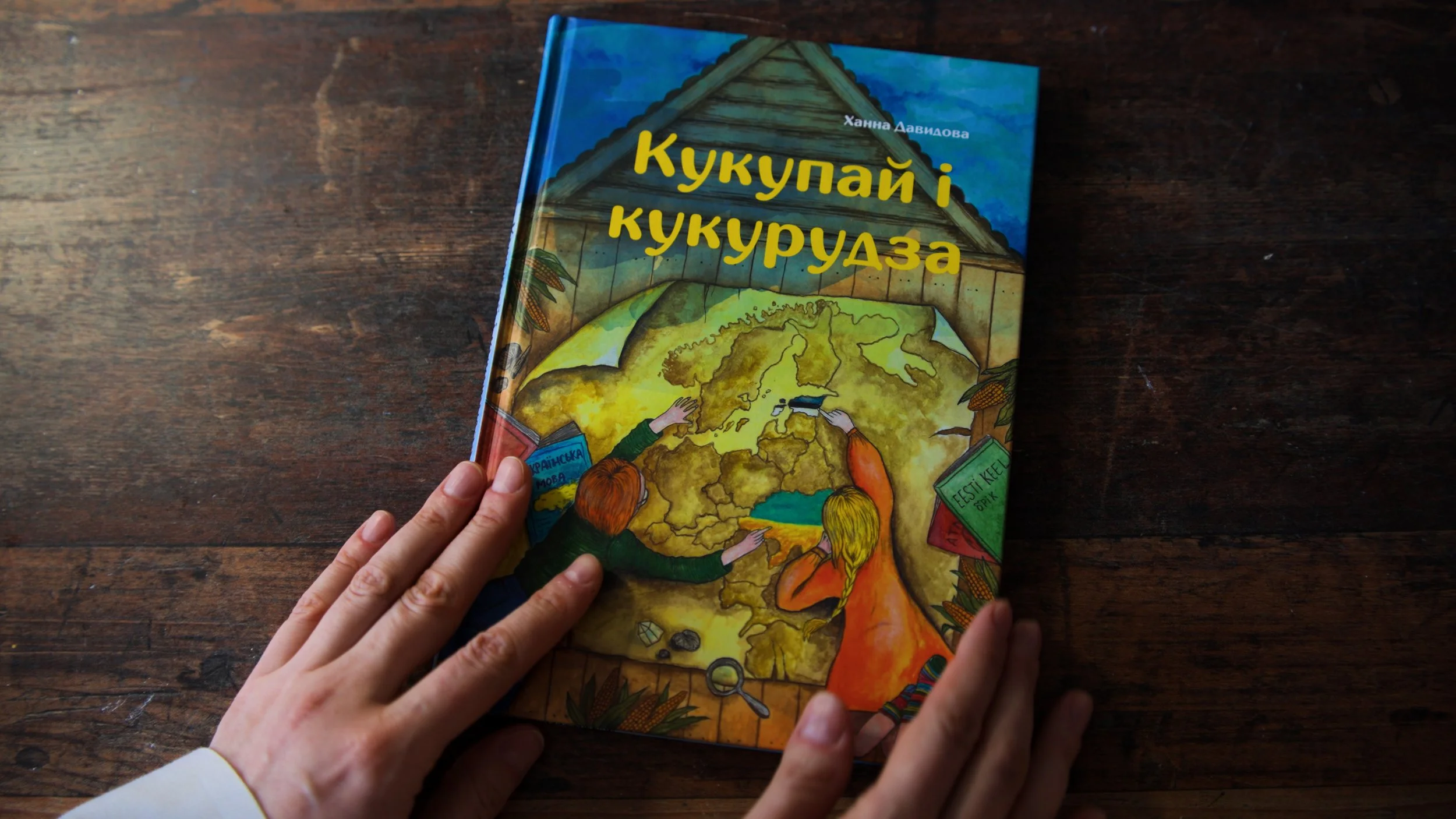 A person with light skin and short nails is holding a colorful children's book titled 'Кукупай і кукурудза'. The book cover shows an illustration of two children pointing at a map of Europe, with drawings of books, plants, and a magnifying glass around them, on a dark wooden surface.
