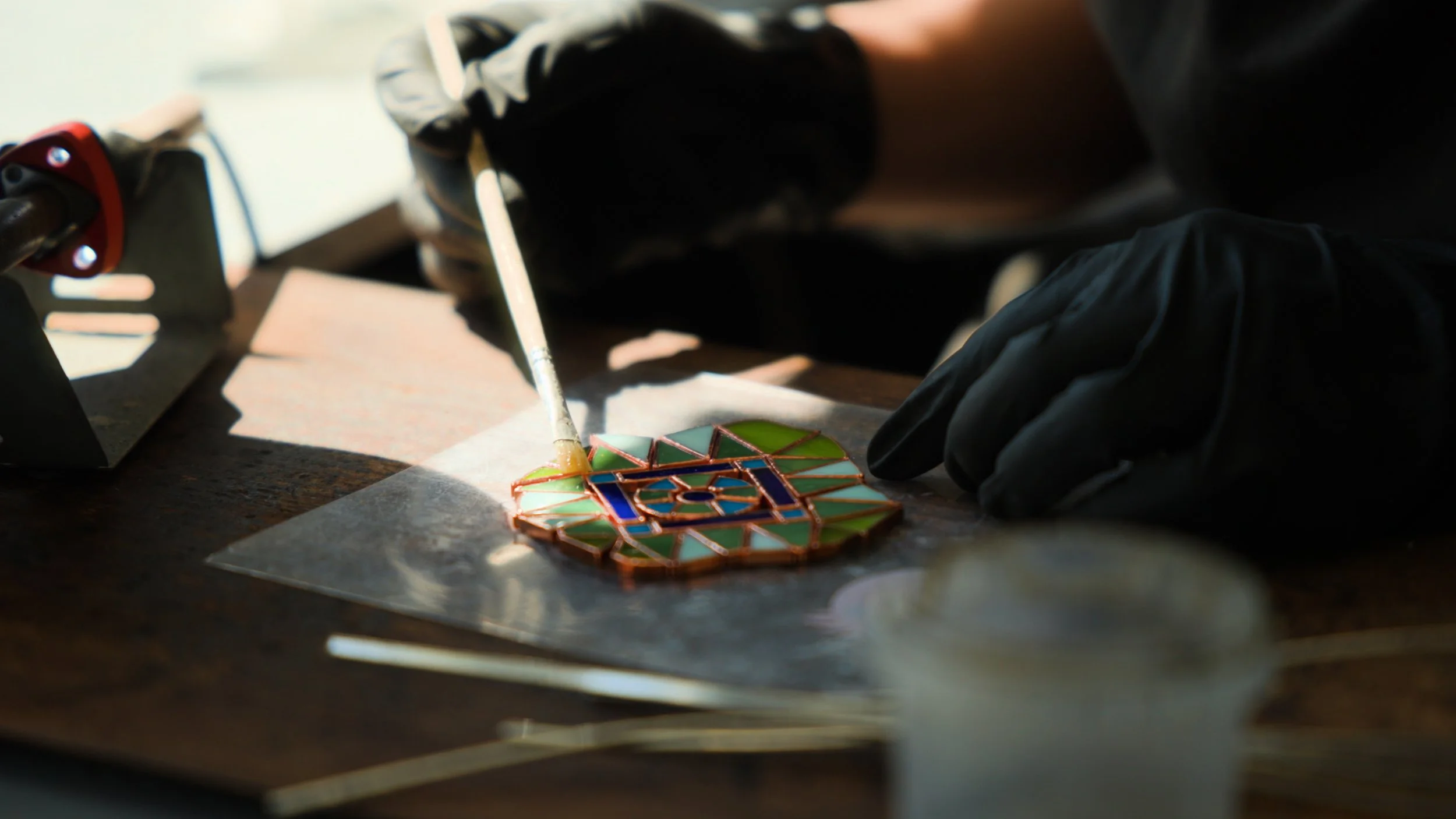 A person is creating a stained glass mosaic, applying yellow paint with a brush onto the colored glass pieces arranged on a work surface. The person is wearing black gloves and a dark shirt. Various tools and materials are scattered around.