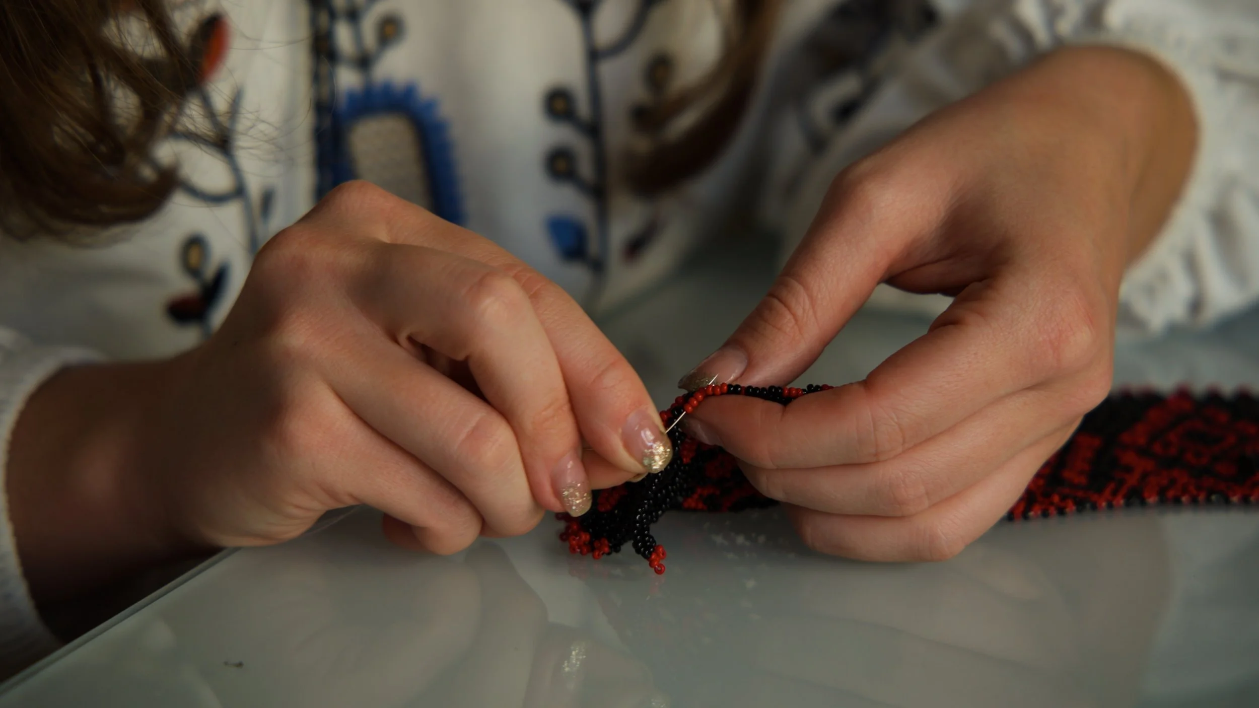 Close-up of hands beading a black and red thread necklace on a white surface.