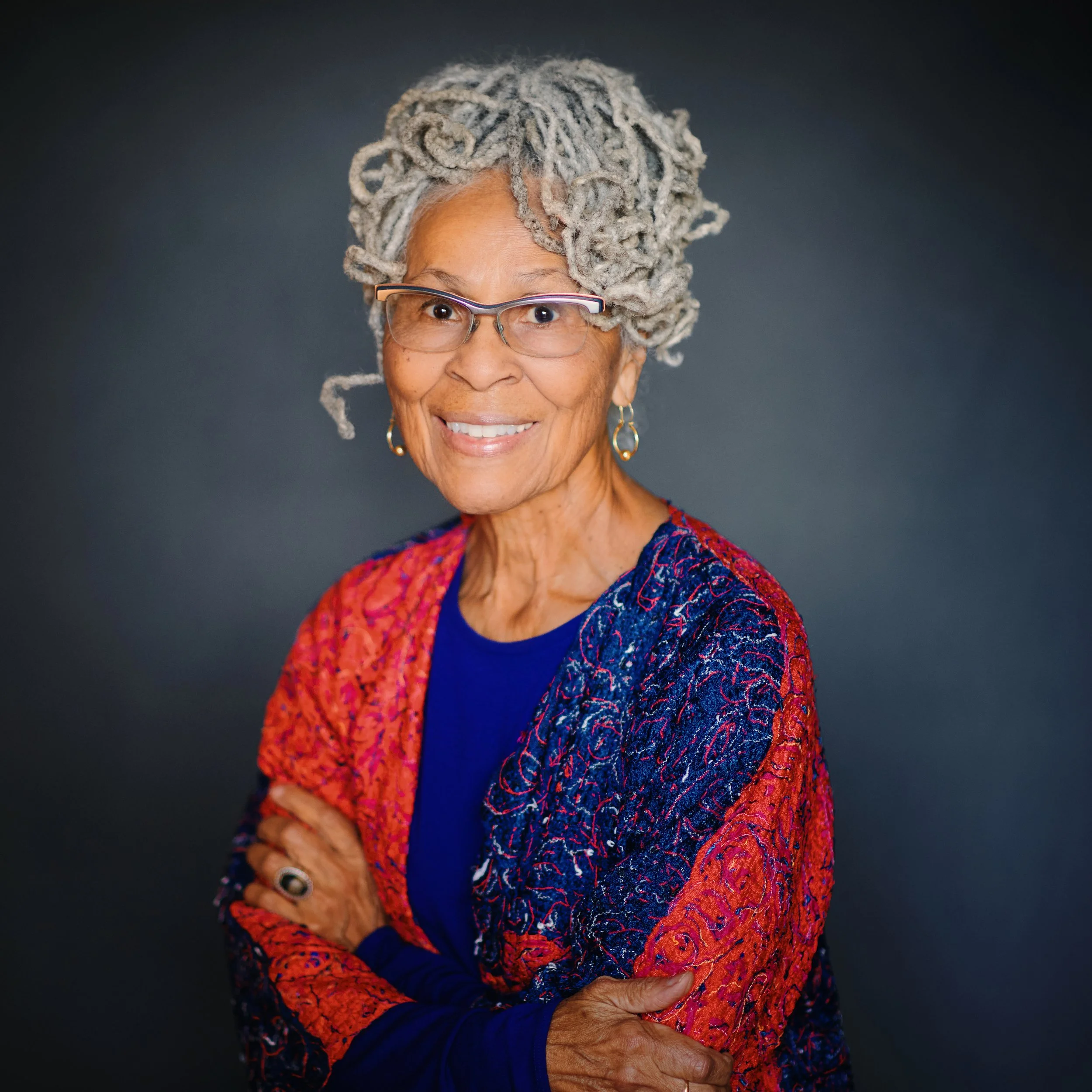 Portrait of an elderly woman with gray dreadlocks, wearing glasses, gold hoop earrings, a colorful red and blue patterned shawl, and a blue top, standing against a dark background.