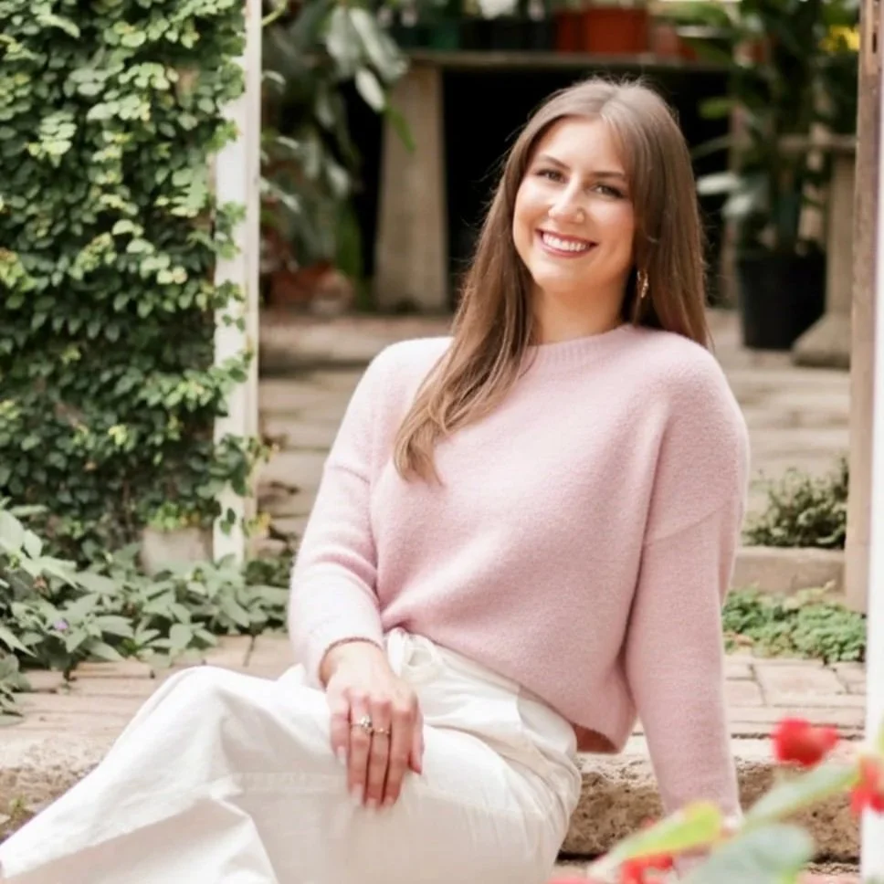 Meredith Mullin, therapist with Can We Meet Therapy. A young woman with long brown hair, smiling and sitting outdoors on a garden path, wearing a light pink sweater and white pants.