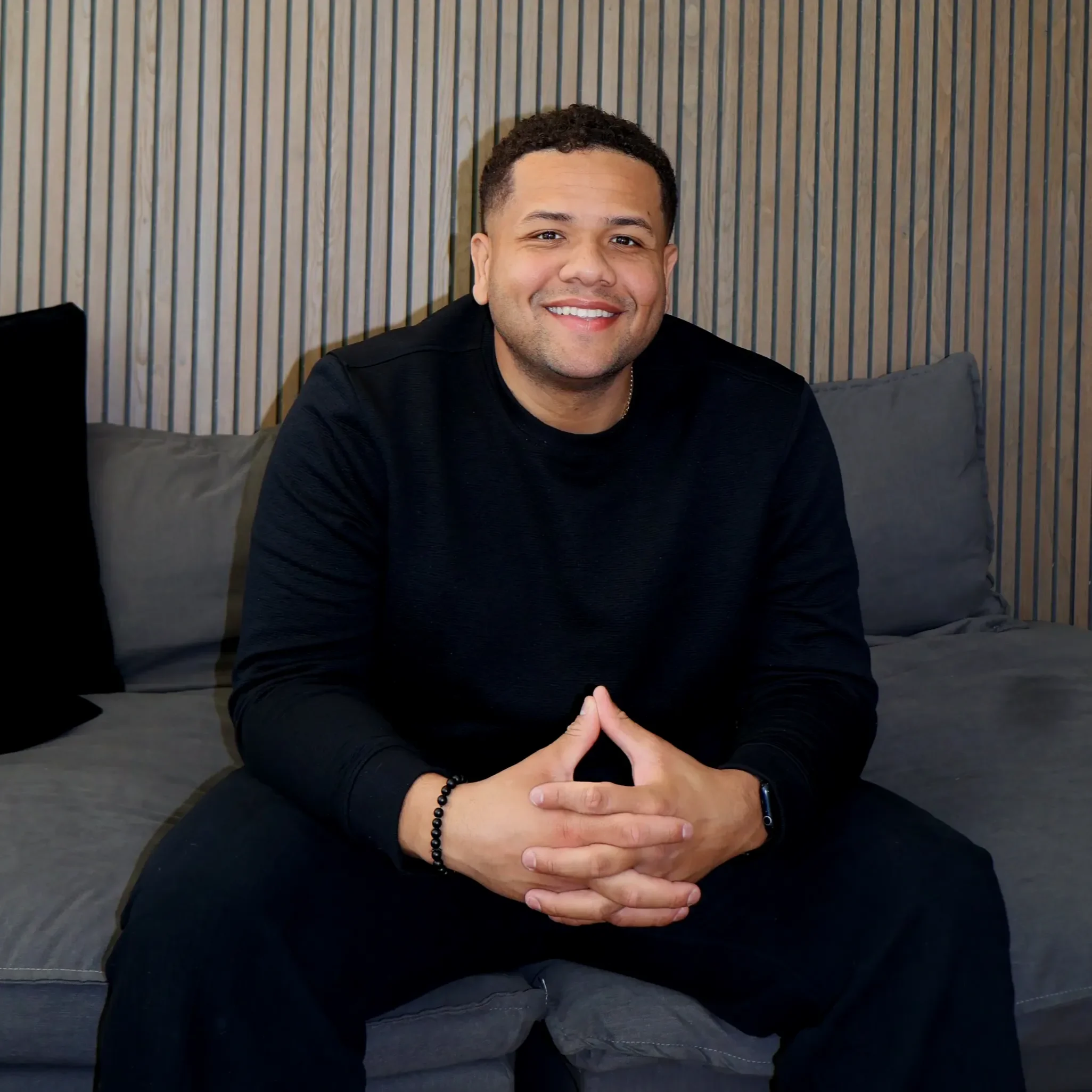 Andres Paz-Colon, podcast host at Can We Talk. Picture of a smiling man with short curly hair sitting on a gray couch with a wooden slat wall background, wearing a black shirt and a black beaded bracelet, with his hands clasped in front of him.