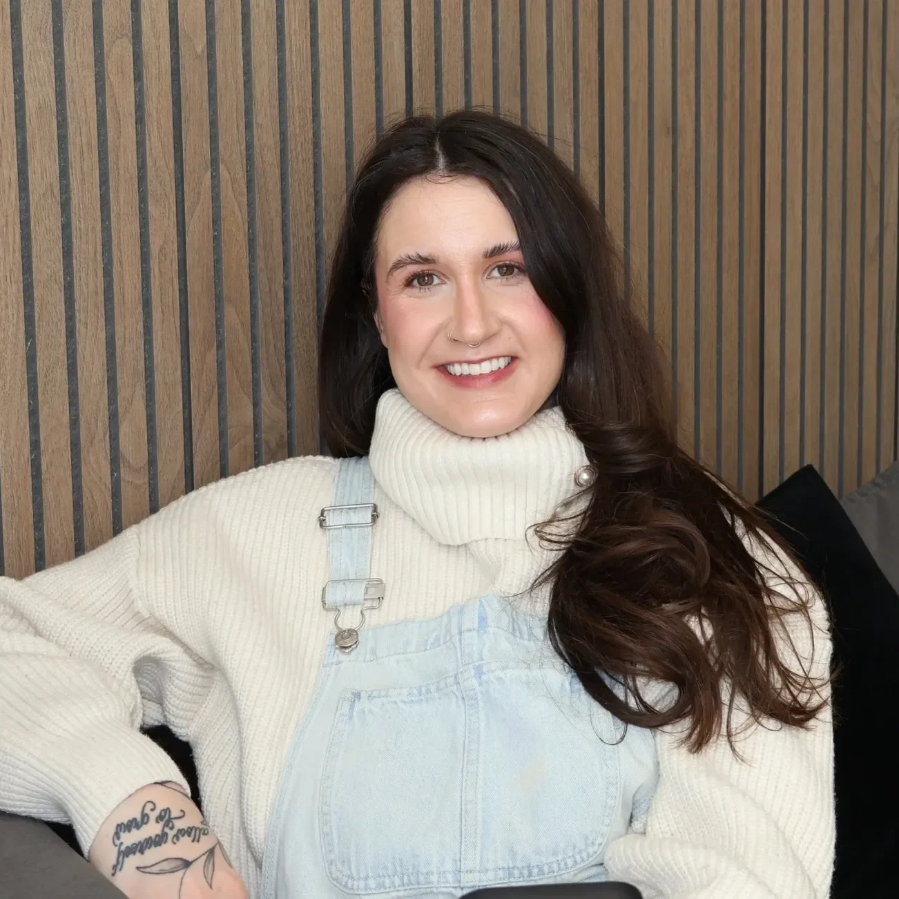 Kelsie Johnson, LMFT therapist with Can We Meet Therapy. Picture of a woman with long dark hair smiling, wearing a cream turtleneck sweater and light blue overalls, sitting in front of a wooden-paneled wall.