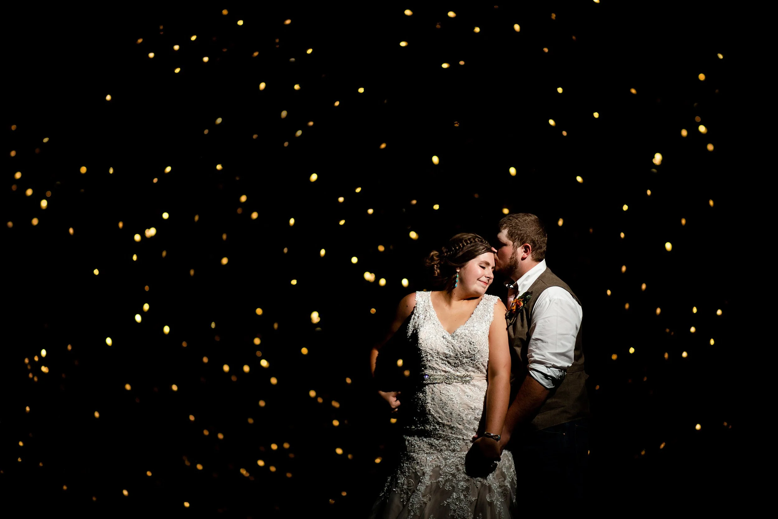 A bride and groom sharing an intimate moment with the groom kissing the bride's forehead against a dark background with floating golden lights.