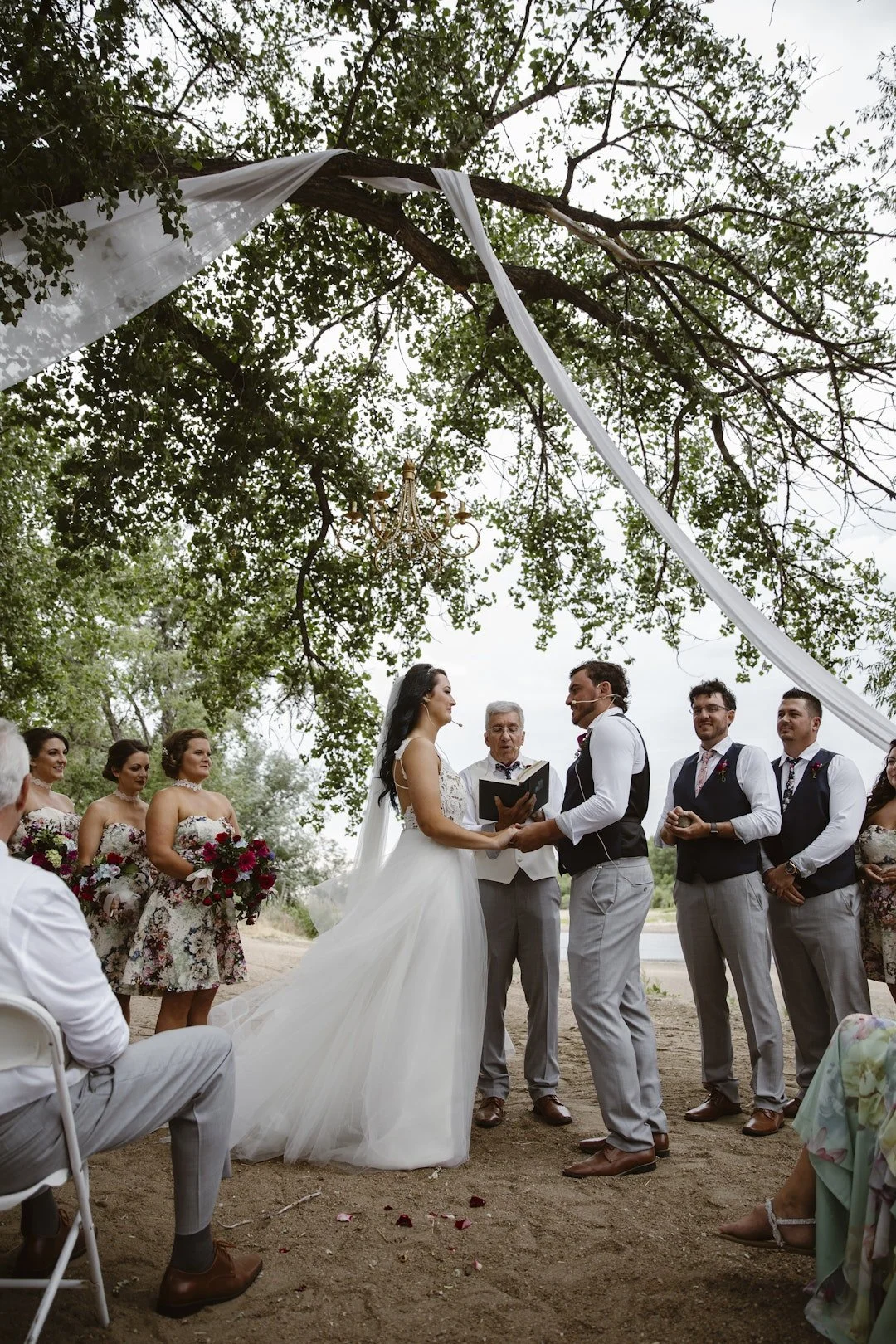 A Colorado outdoor wedding venue space along the river during a wedding ceremony feature the bride, groom and wedding officiant with their bridal parties on either side under a large canopy of beautiful trees
