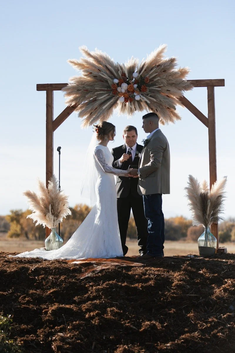 Bride, groom and wedding officiant stand under a rustic arch with wooden beams and pampas grass flower arrangement above them at an outdoor wedding venue featuring a bluff overlooking the countryside