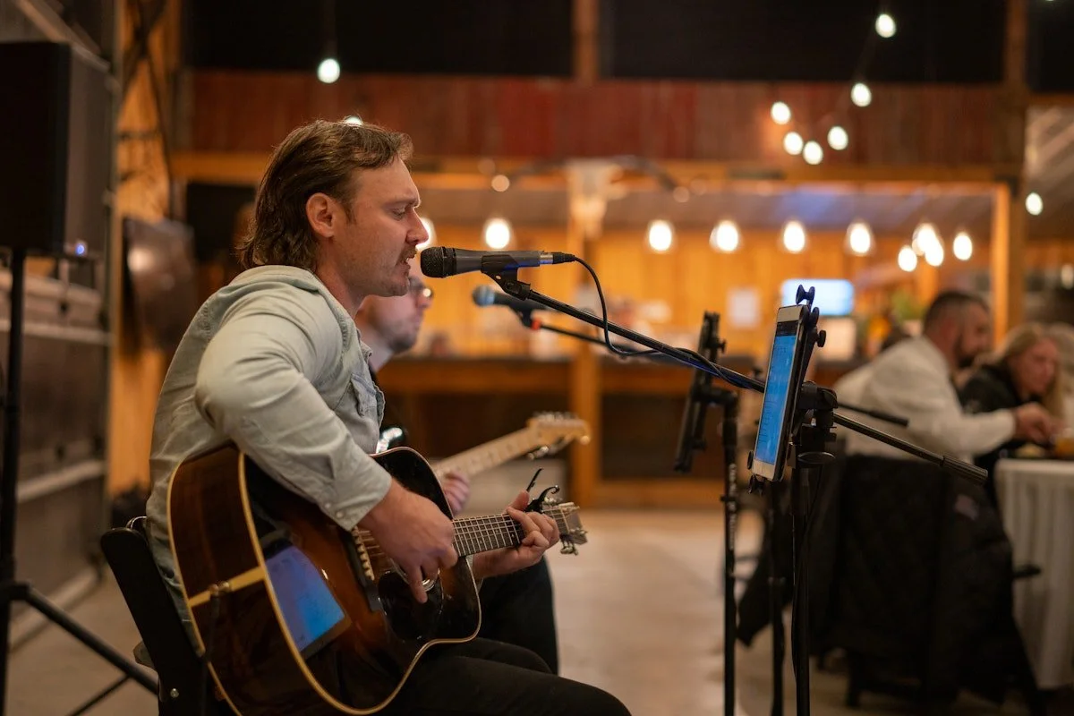 A man with shoulder-length hair playing an acoustic guitar and singing into a microphone at a cozy indoor venue with warm lighting and wooden decor.