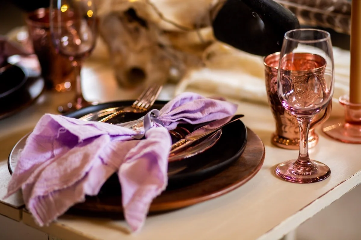A table setting with black plates, pink-tinted water glasses, a purple napkin, and a gold fork and knife, with a decorative skull in the background.