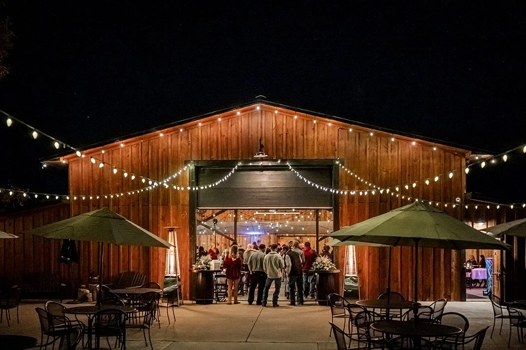 Exterior of a wooden barn private event venue with Edison lights strung across the outdoor event spaces and people gathered inside the barn