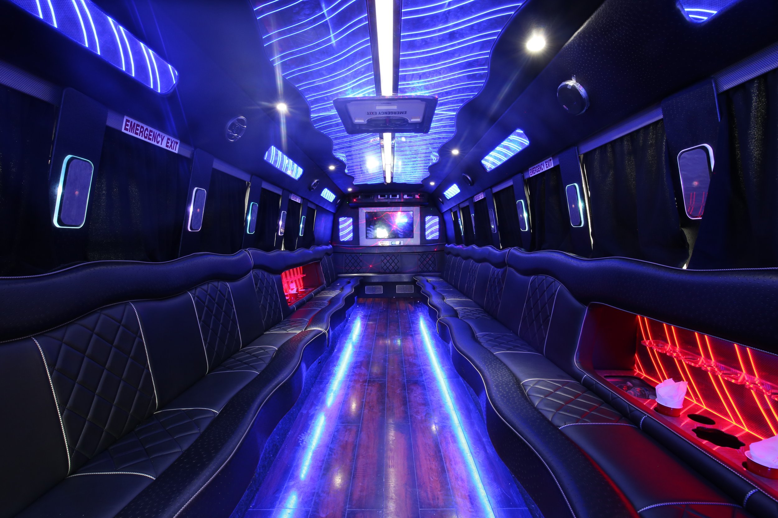 Interior of a luxury limousine with black leather seating along both sides, illuminated by blue and red LED lights, with a television screen at the front and emergency exit signs on both sides.