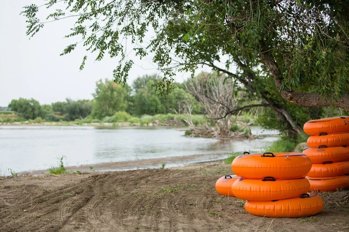 Stacked orange inner tubes on a sandy riverbank under a tree overlooking a calm river with green trees in the background.