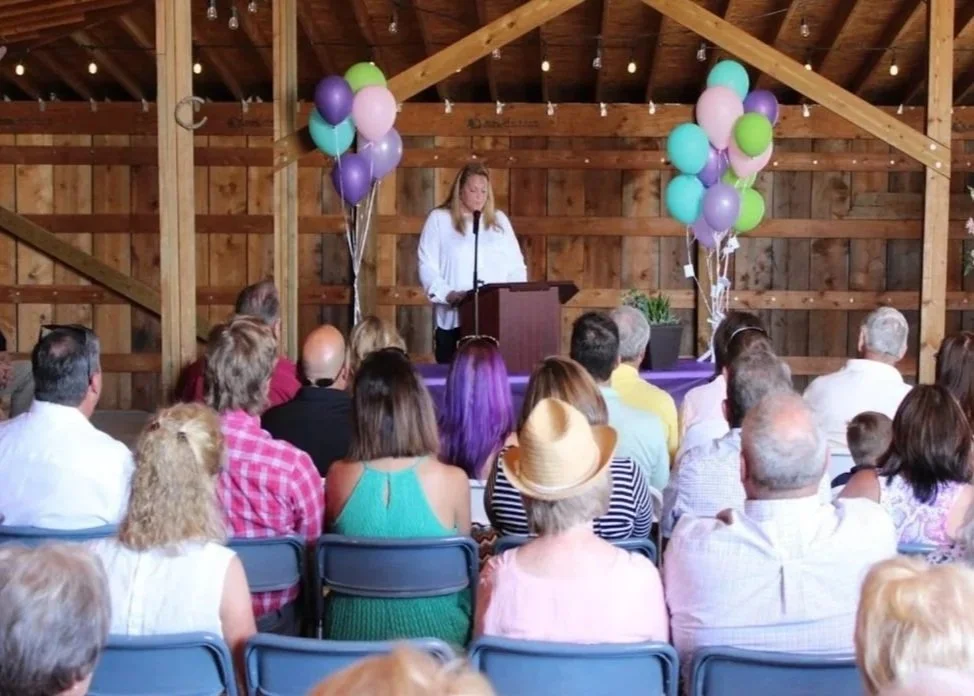 Wooden barn corporate event venue filled with seated people looking at a speaker on a podium with balloons on either side