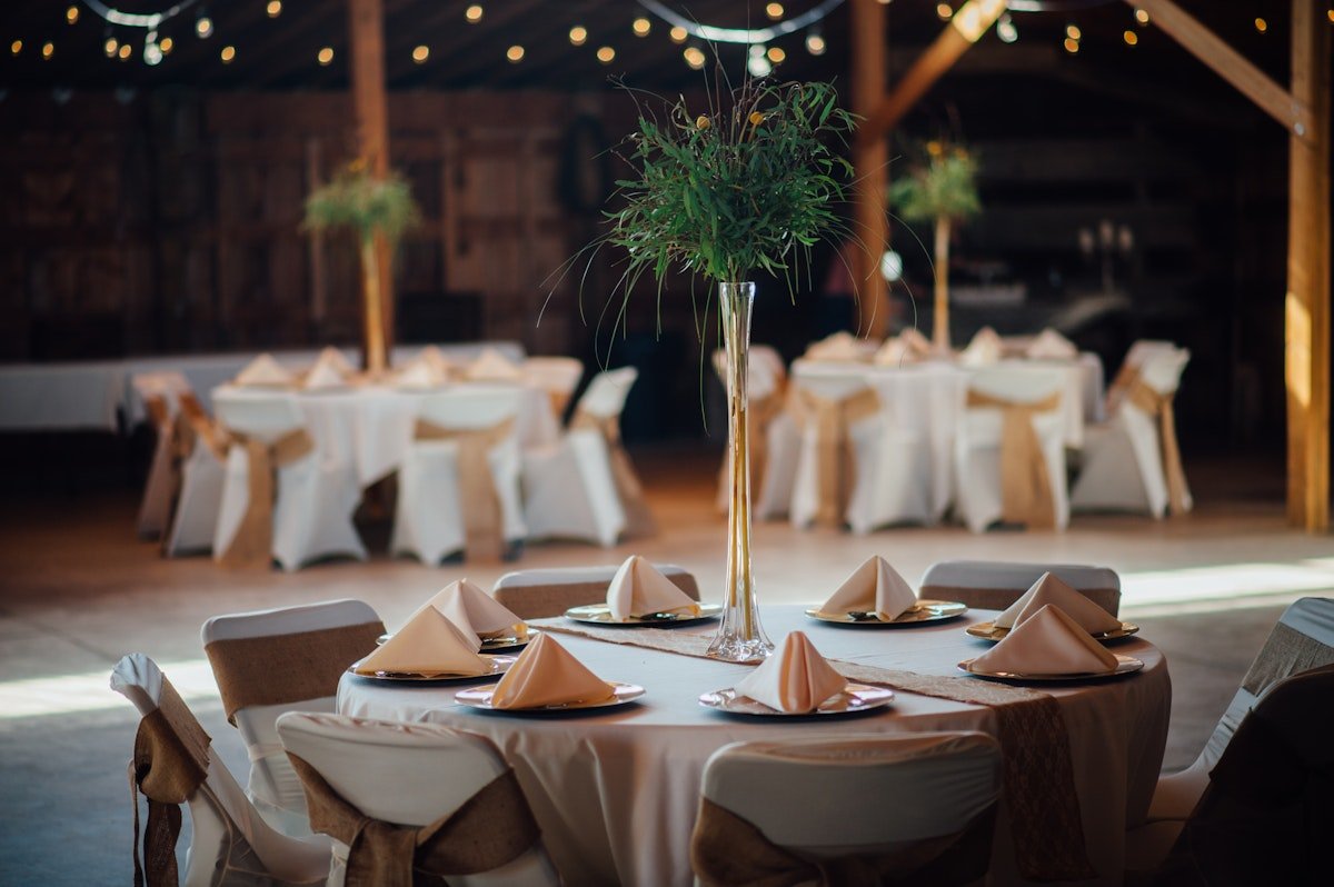 Wedding reception with round tables draped in white linens, decorated with folded napkins on plates, and centerpieces consisting of tall, slender vases with greenery. The room has wooden walls and beams, with soft lighting and a rustic ambiance.