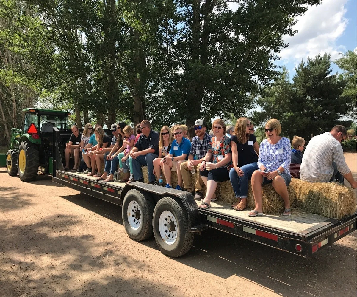 A group of people sitting on hay bales on a flatbed trailer, attached to a tractor, in an outdoor setting with trees and blue sky.