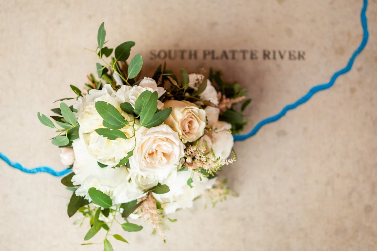 A floral arrangement of white and pale blush roses, white hydrangeas, green leaves, and small pink filler flowers on a map background showing the South Platte River.
