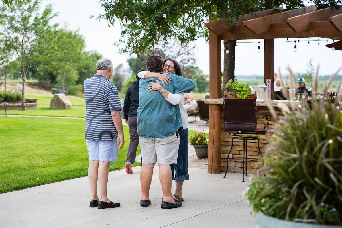 People hugging outdoors at a gathering or event, with a grassy area and trees in the background.
