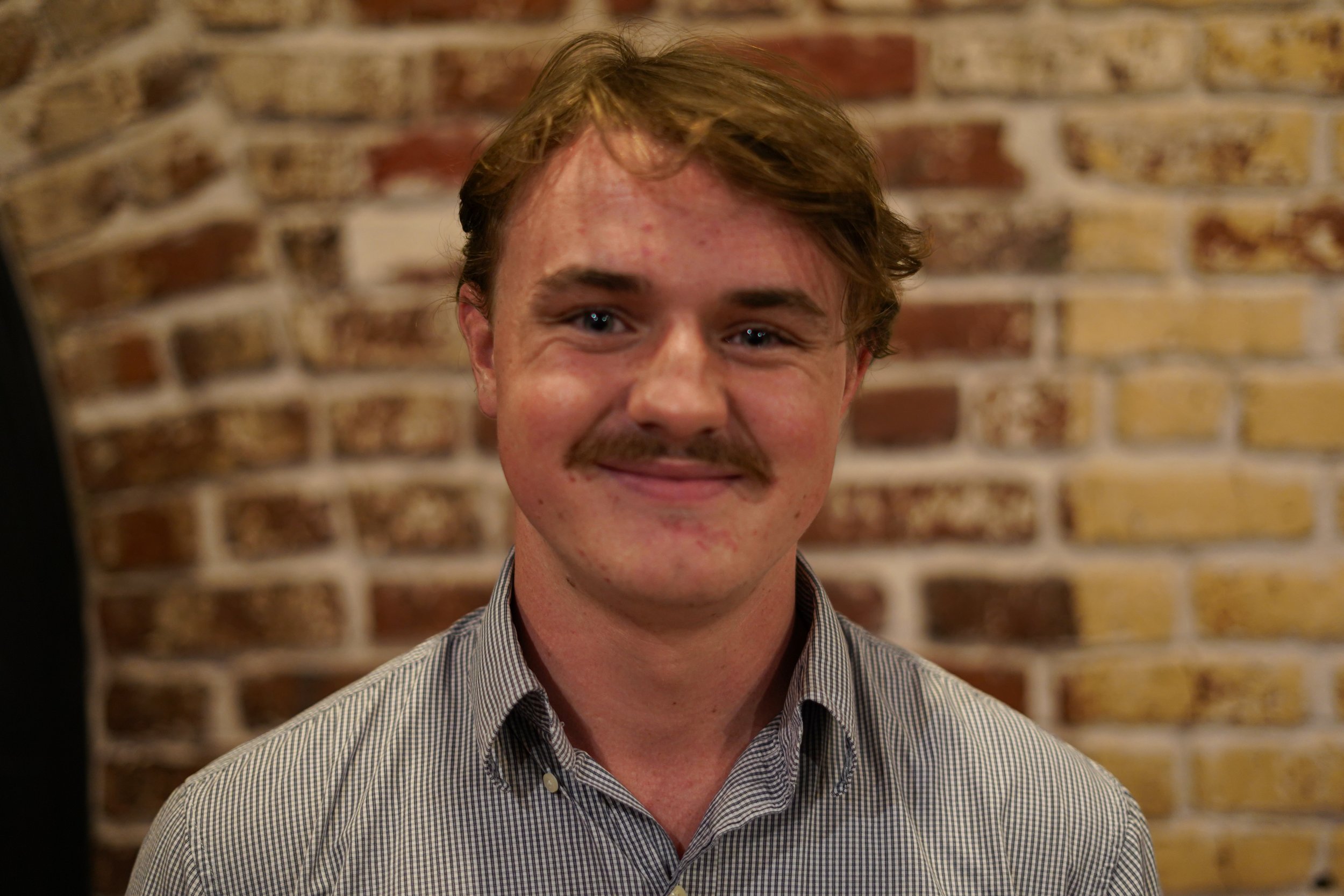 A young man with light brown hair, blue eyes, a mustache, and a slight smile, wearing a gray checkered shirt, standing in front of a brick wall.