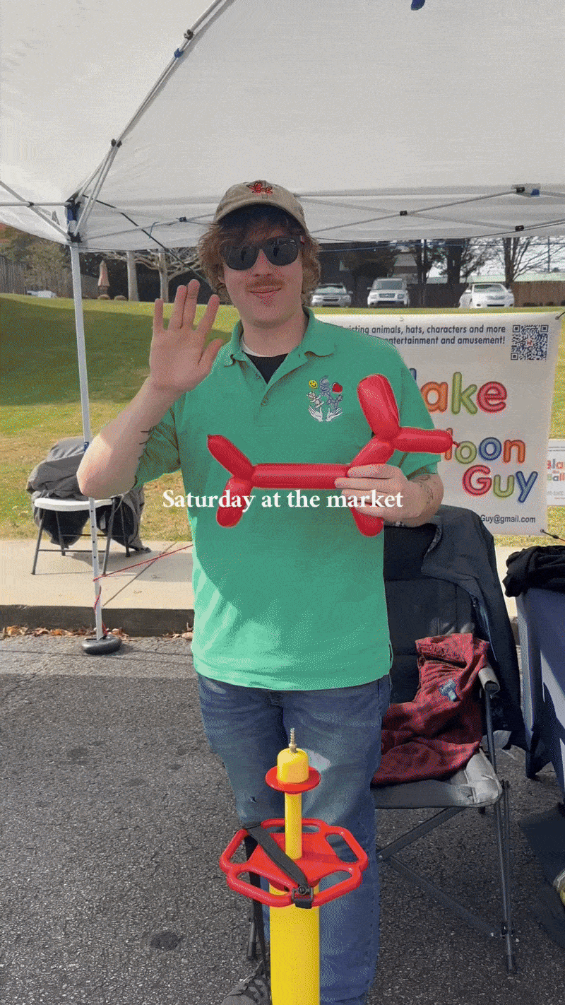 A young man wearing sunglasses and a green polo shirt holding a red balloon dog sculpture, standing at an outdoor market on a cloudy day under a white canopy.