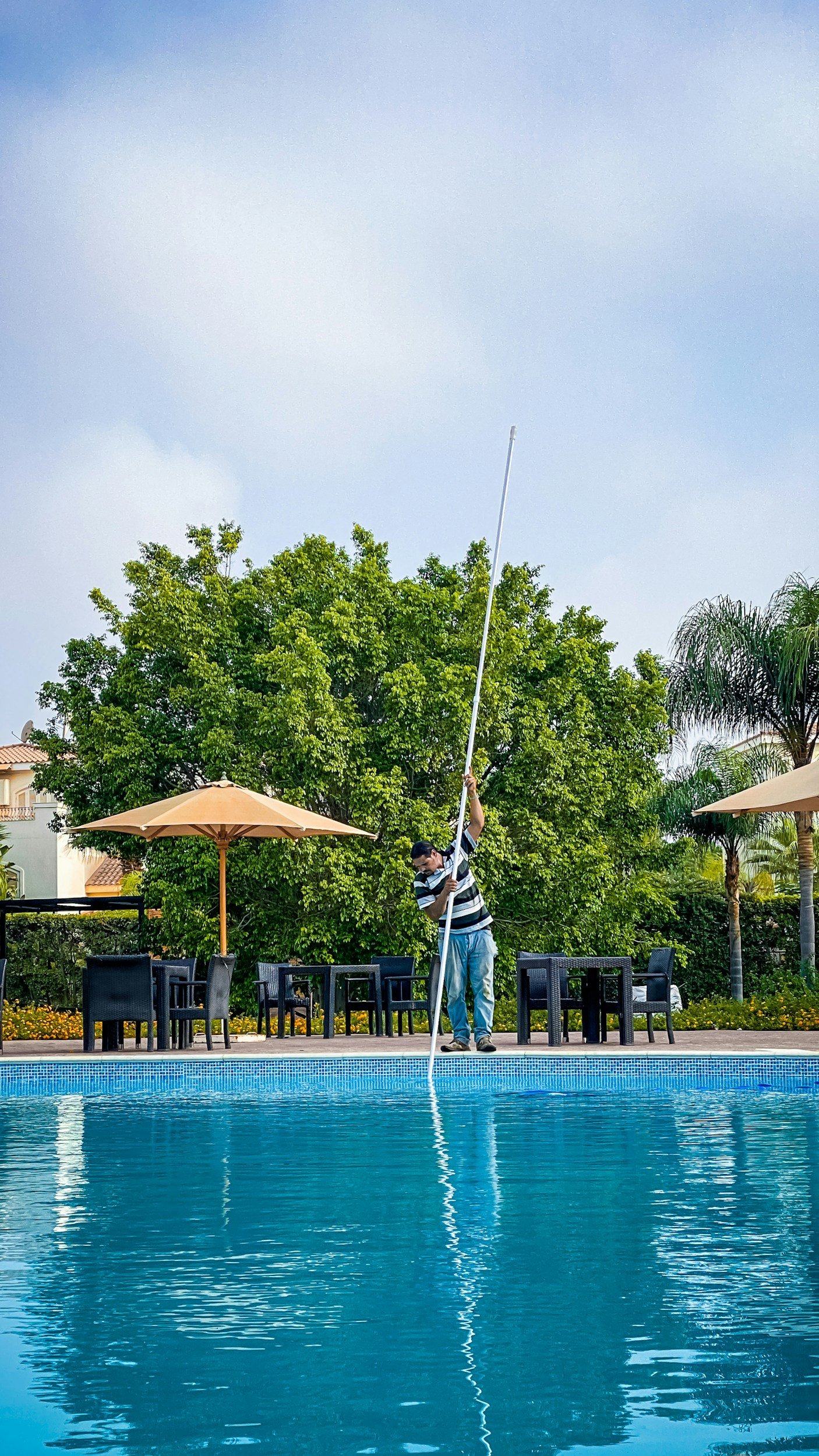 Person setting up a long pole near a swimming pool with outdoor chairs, umbrellas, trees, and buildings in the background.