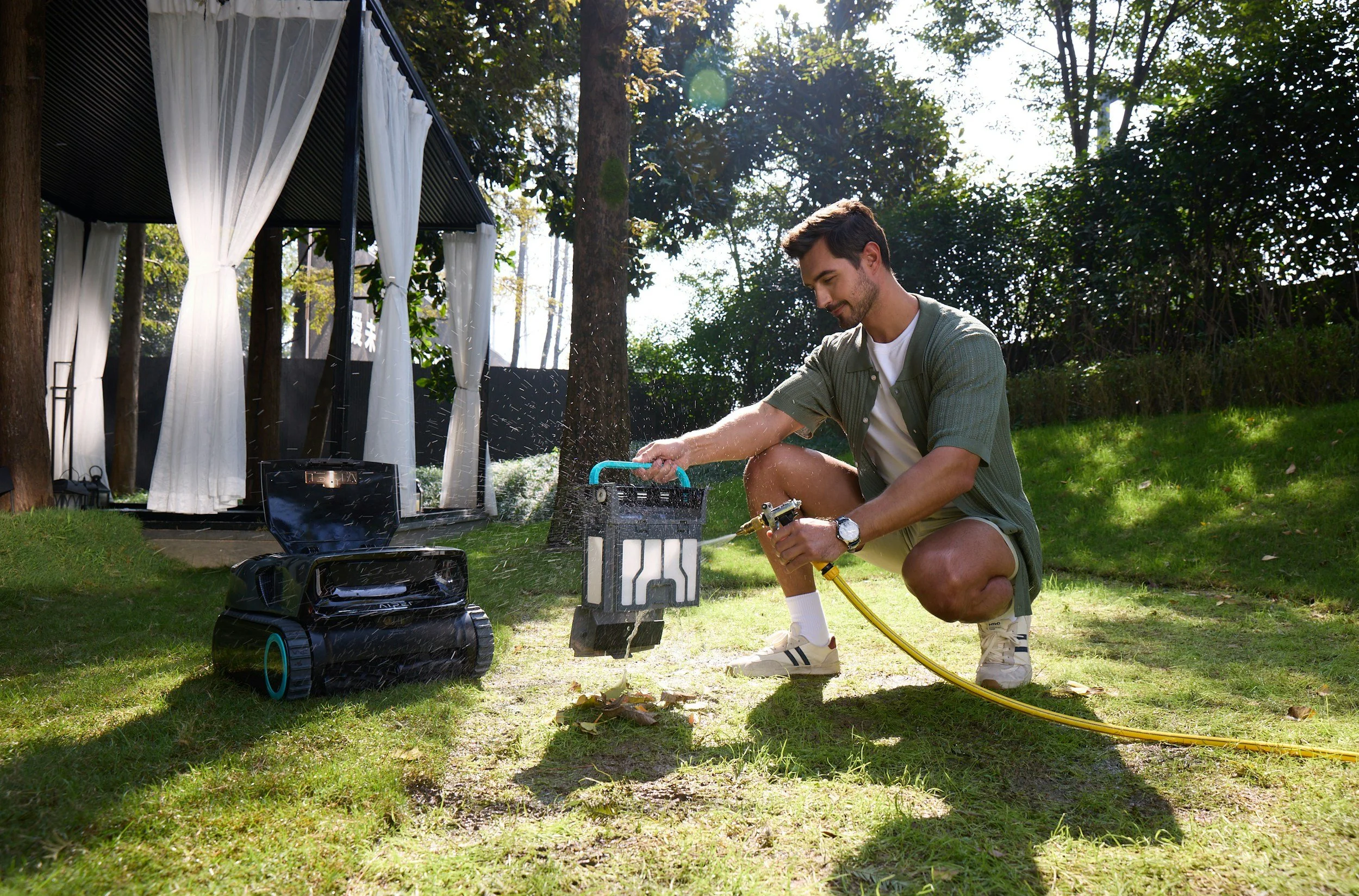 A man kneeling on the grass outdoors, watering plants with a garden hose. There is a black robot vacuum on the grass nearby, and a stand with a scissors and other tools. In the background, there are trees, a small structure with white curtains, and sunlight filtering through the leaves.