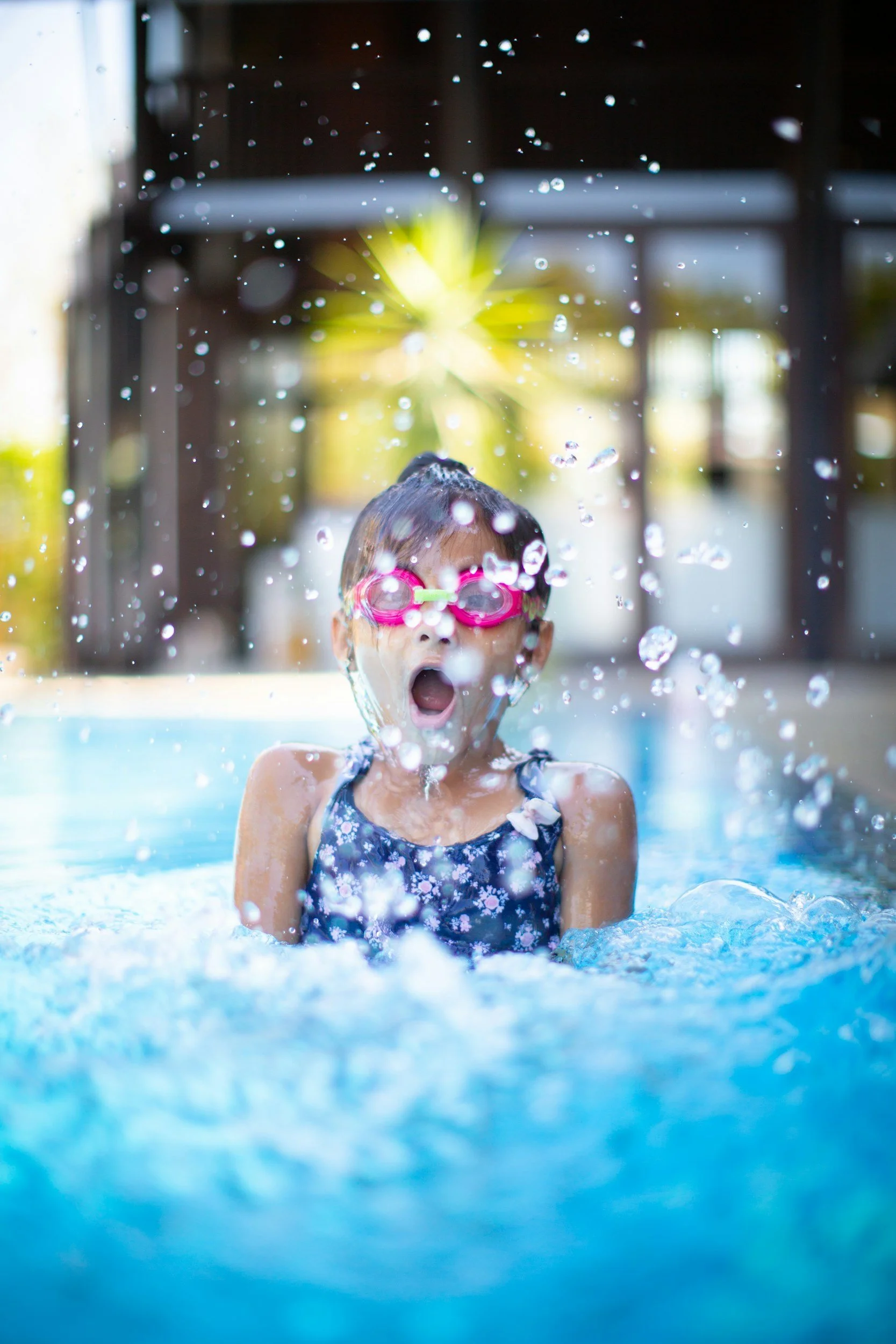 A young girl wearing pink goggles and a blue floral swimsuit is playing in a swimming pool, with water splashing around her and her mouth open in excitement.
