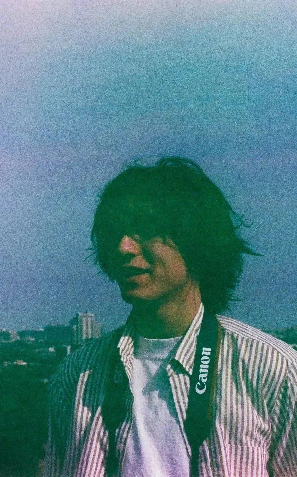 Cullen Thurston with shaggy, dark hair standing outdoors at dusk, wearing a striped shirt and a Canon camera strap around their neck, with a city skyline in the background.