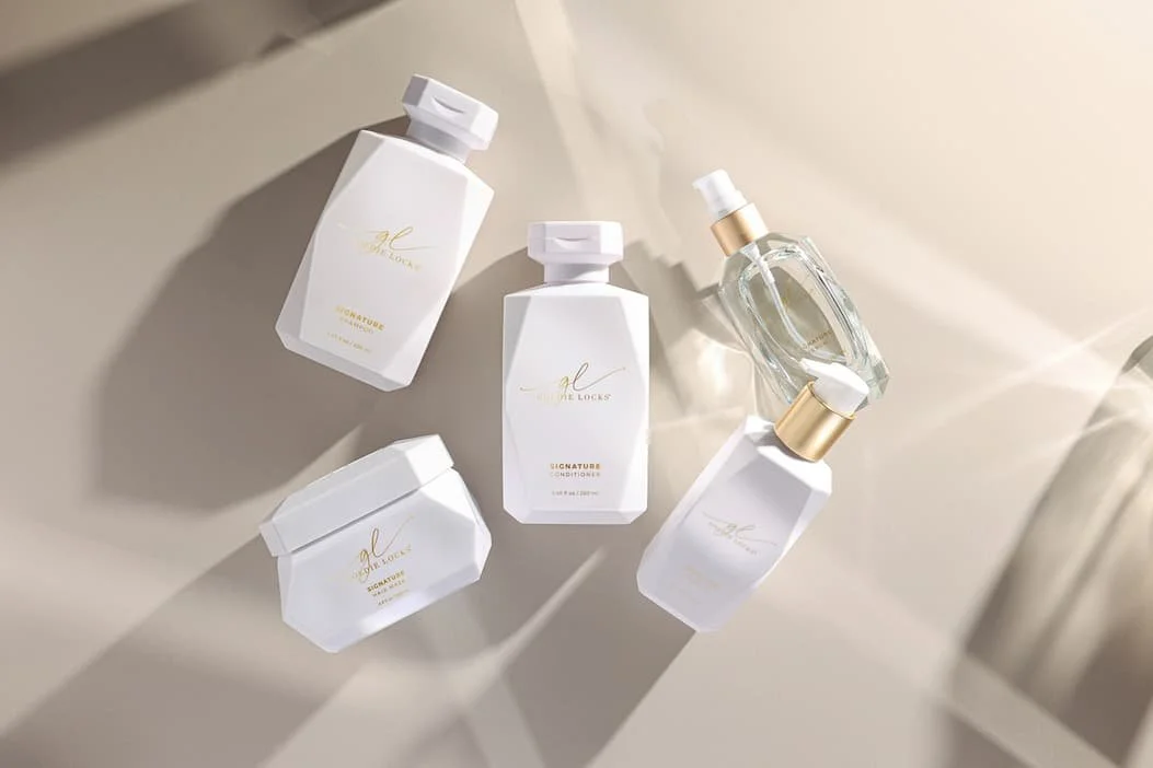 Collection of white bottles and containers with gold accents, including a spray bottle with a clear liquid, on a light-colored surface.