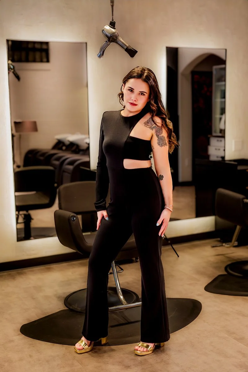 A woman with long brown hair and tattoos on her arms is standing in a salon or barbershop, wearing a black outfit with cutouts. A hairdryer is hanging from the ceiling above her.