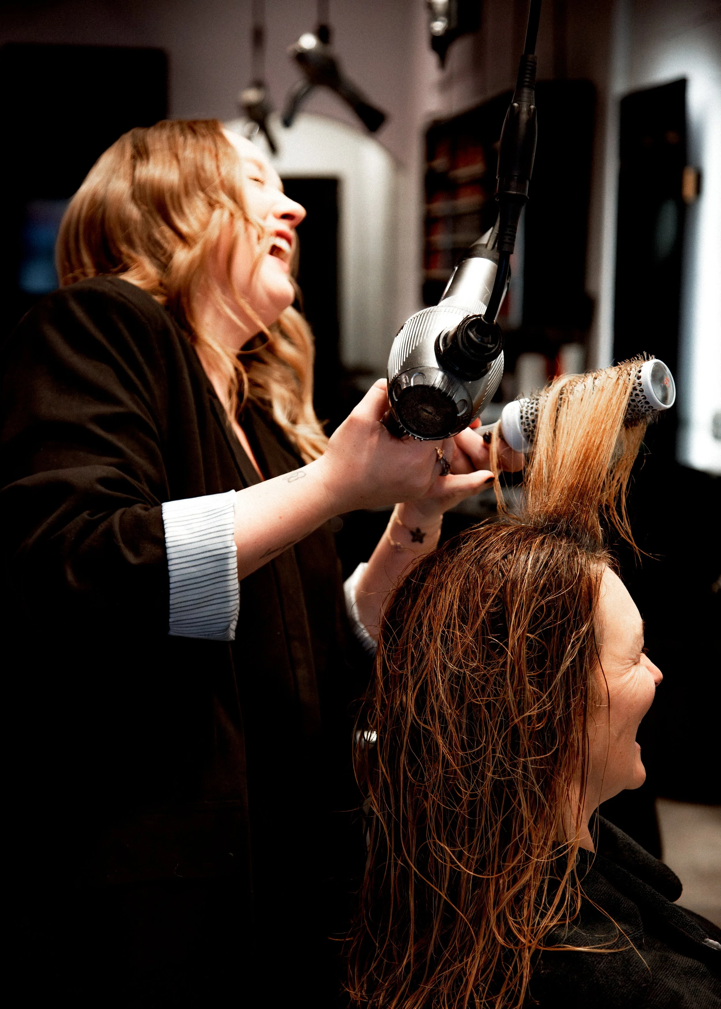 A woman gets her red hair styled with a blow dryer and curling brush at a salon, laughing or smiling.