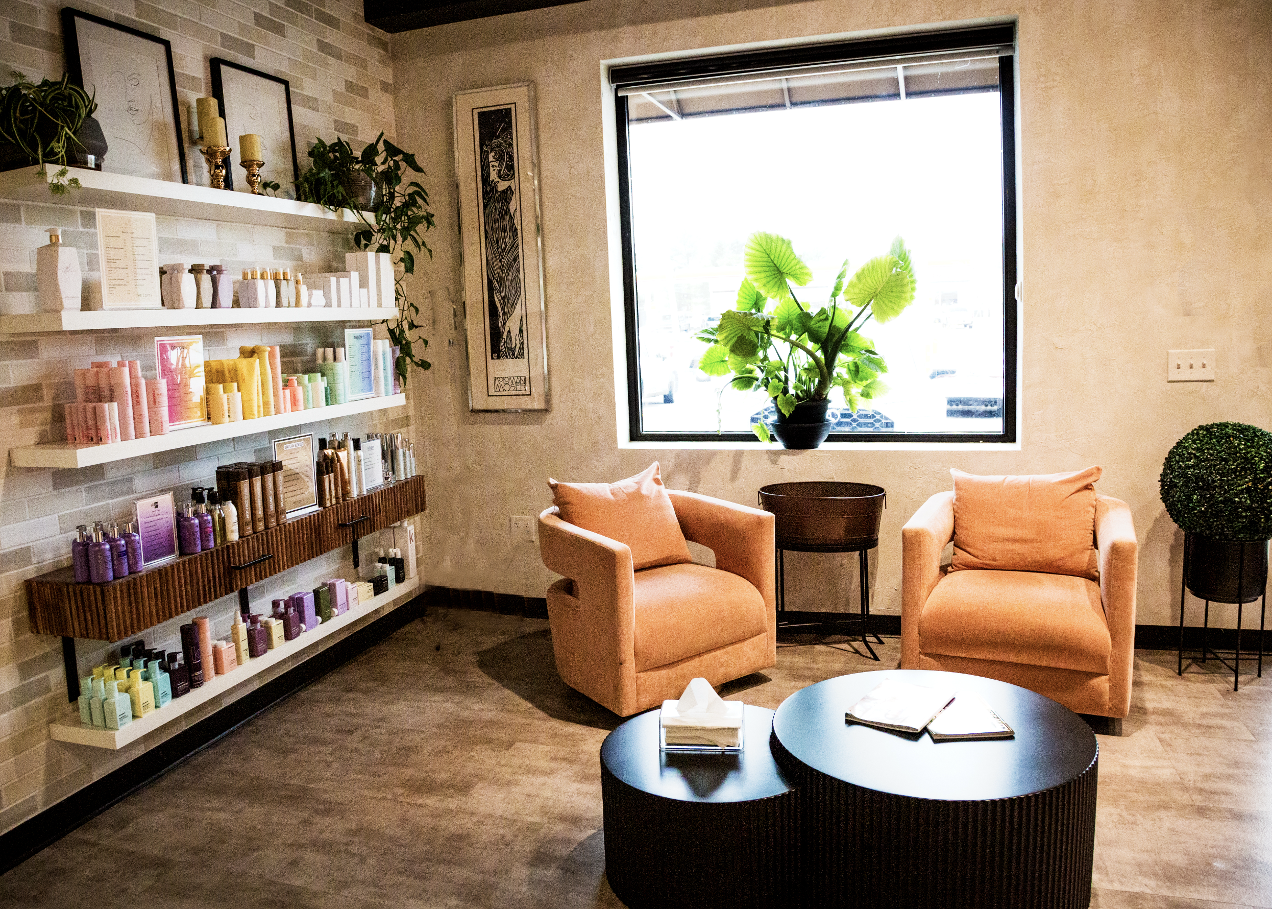 A cozy waiting area with two peach-colored armchairs, a round black coffee table with notebooks and a tissue box, a large window with a potted plant, wall art, and shelves with colorful skincare or beauty products.