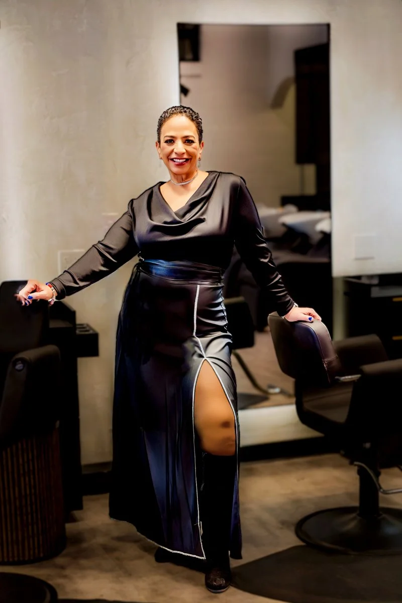 Woman dressed in black, standing in an office with a mirror behind her, smiling at the camera.