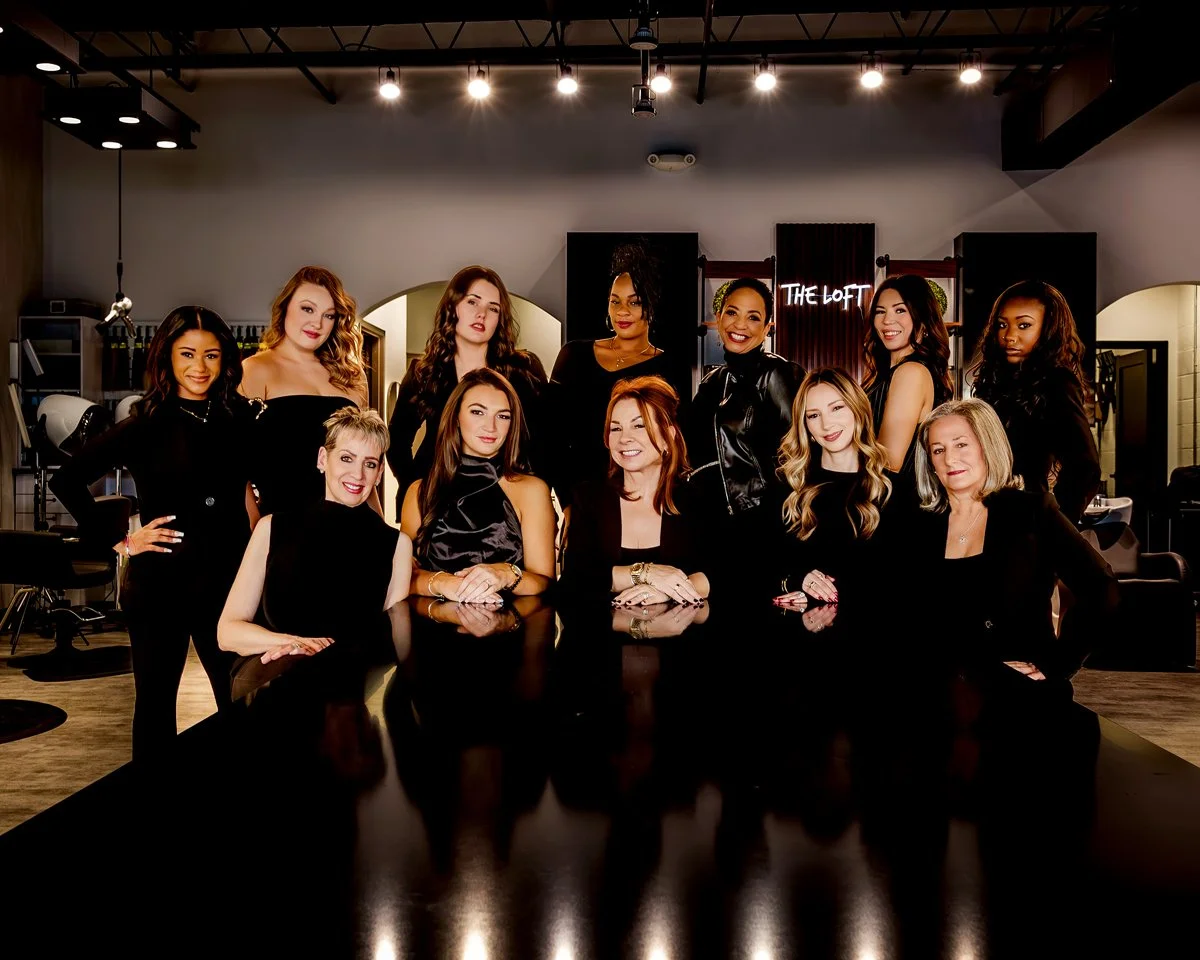 Group of women gathered in a salon or studio, some seated at a black table and others standing behind, with professional hair and makeup, bright lighting, and modern decor in the background.
