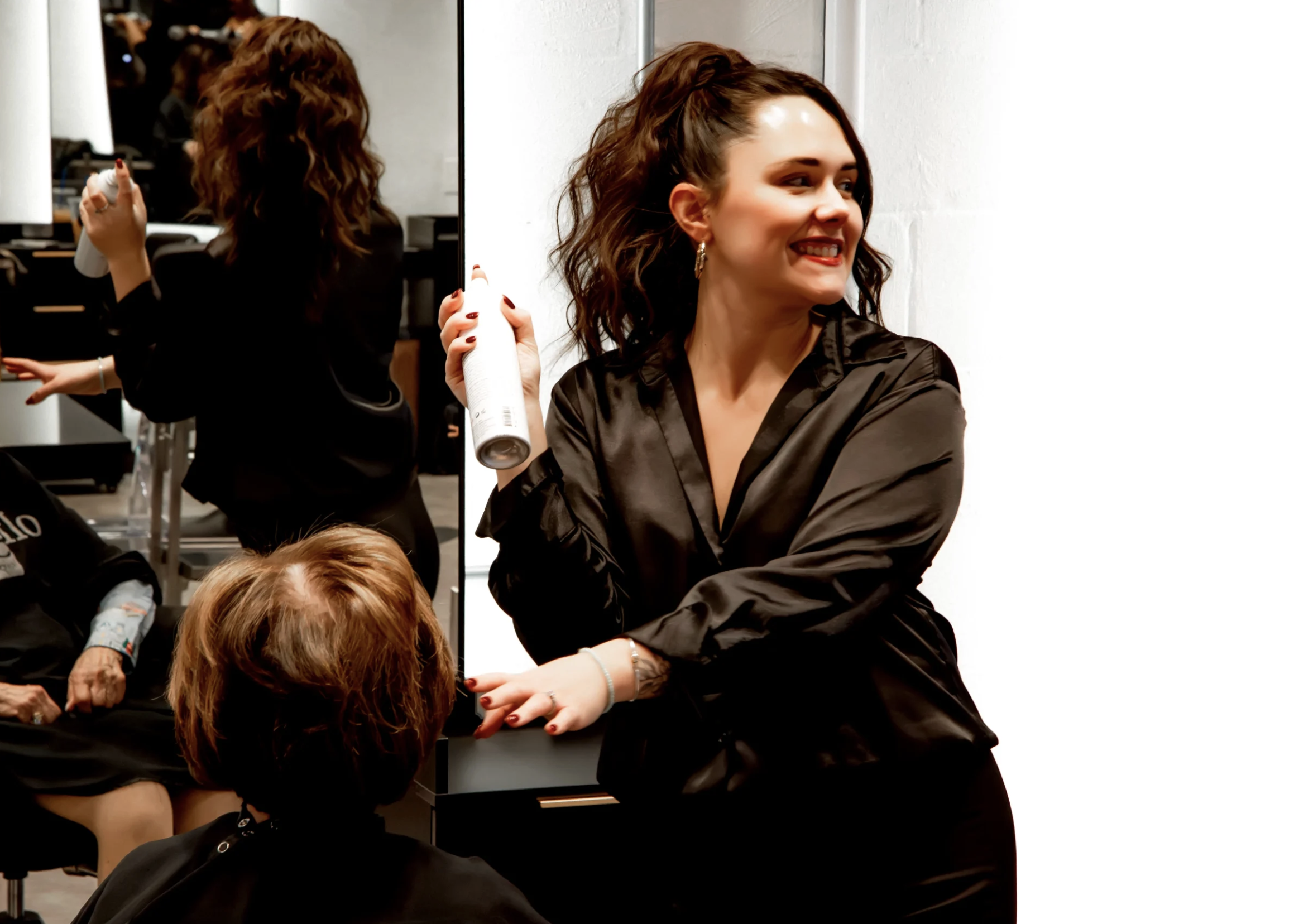 A woman with dark, wavy hair holding a hairspray can, smiling and looking to her right, wearing a black satin robe in a salon or studio.
