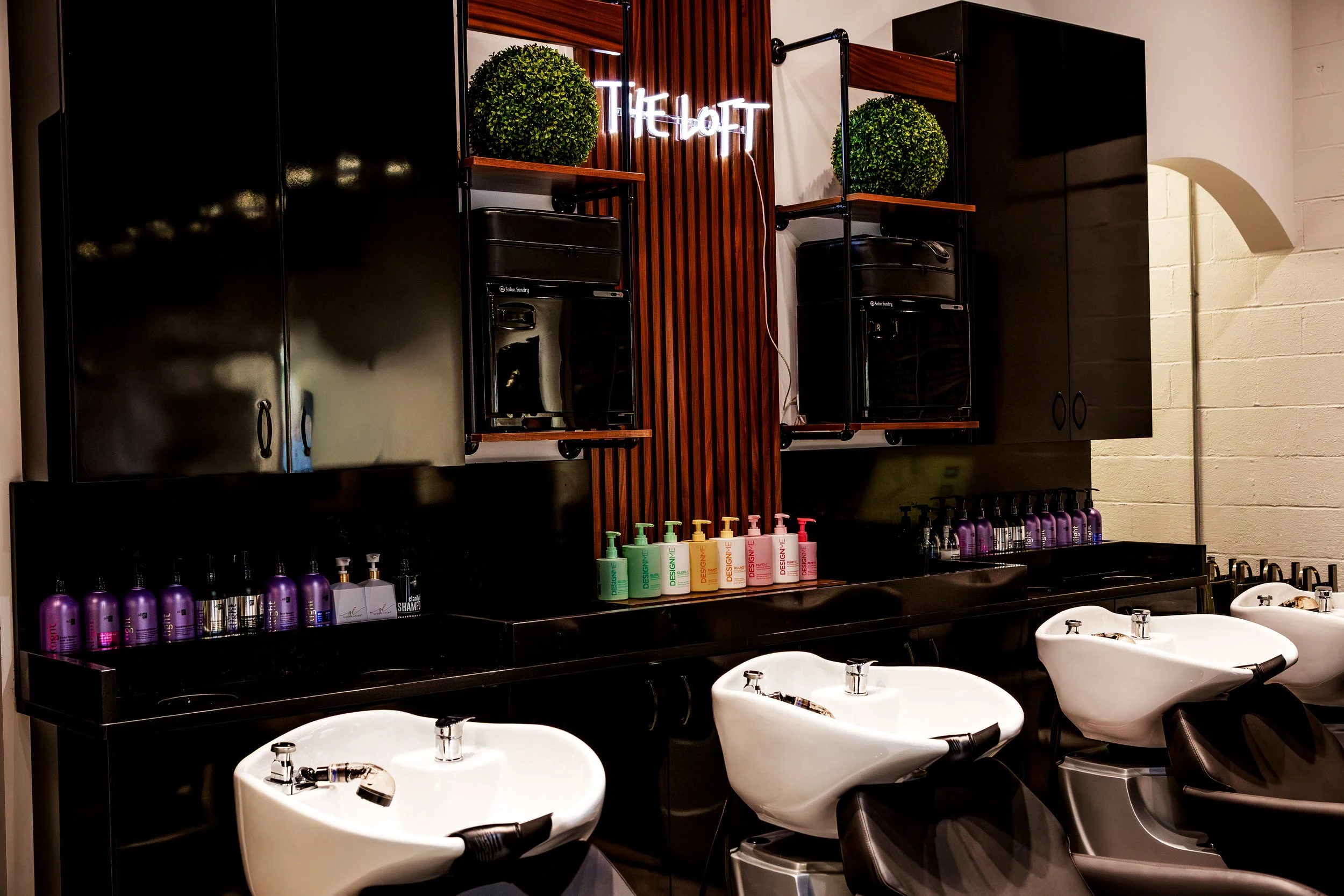 Salon wash stations with white sinks, black countertops, and bottles of hair products, with green plants and a neon sign that says 'The Loft' on a wooden wall in the background.