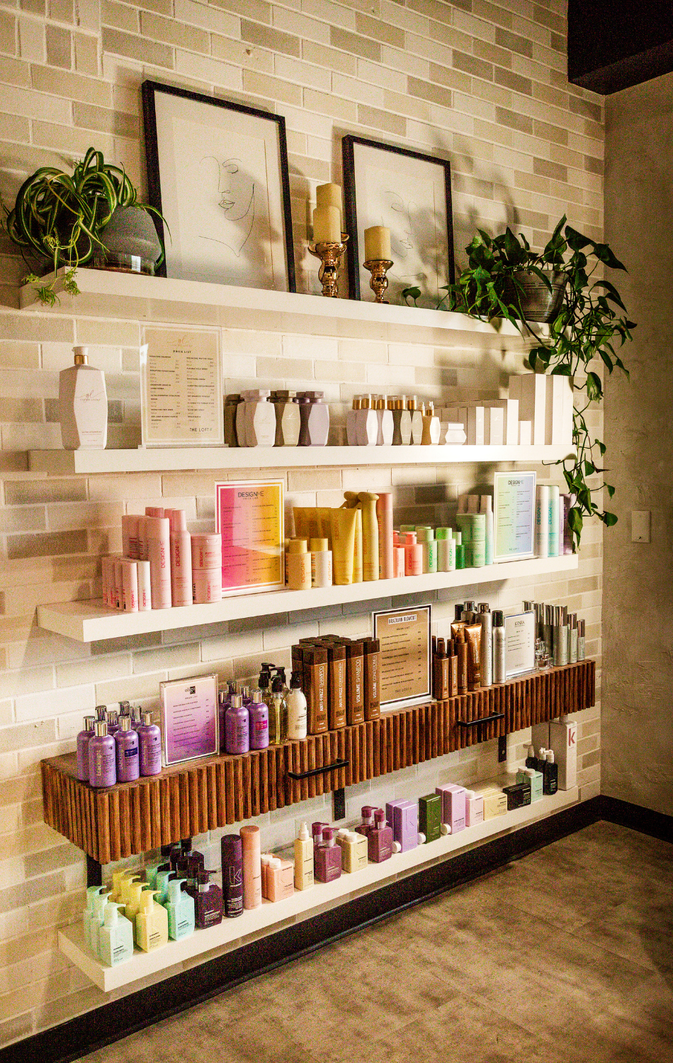 Display of hair and beauty products on white shelves against a brick wall, with framed minimalist line art, potted plants, and decorative candles on the top shelf.